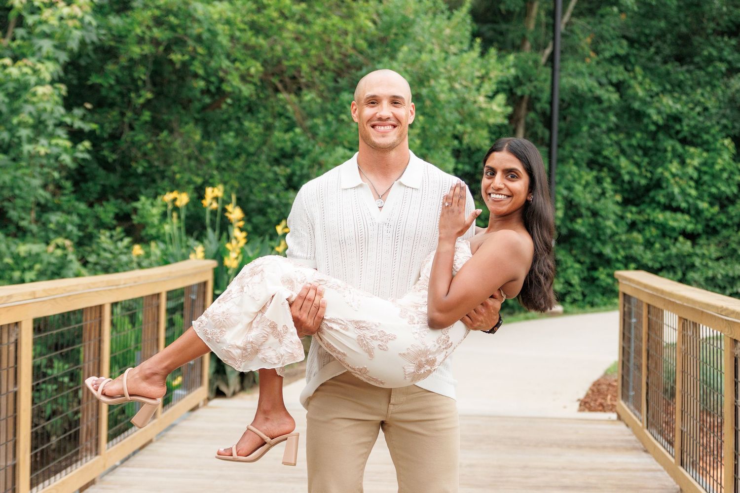 A smiling man in white shirt carries a woman in floral dress on a wooden bridge surrounded by lush greenery.