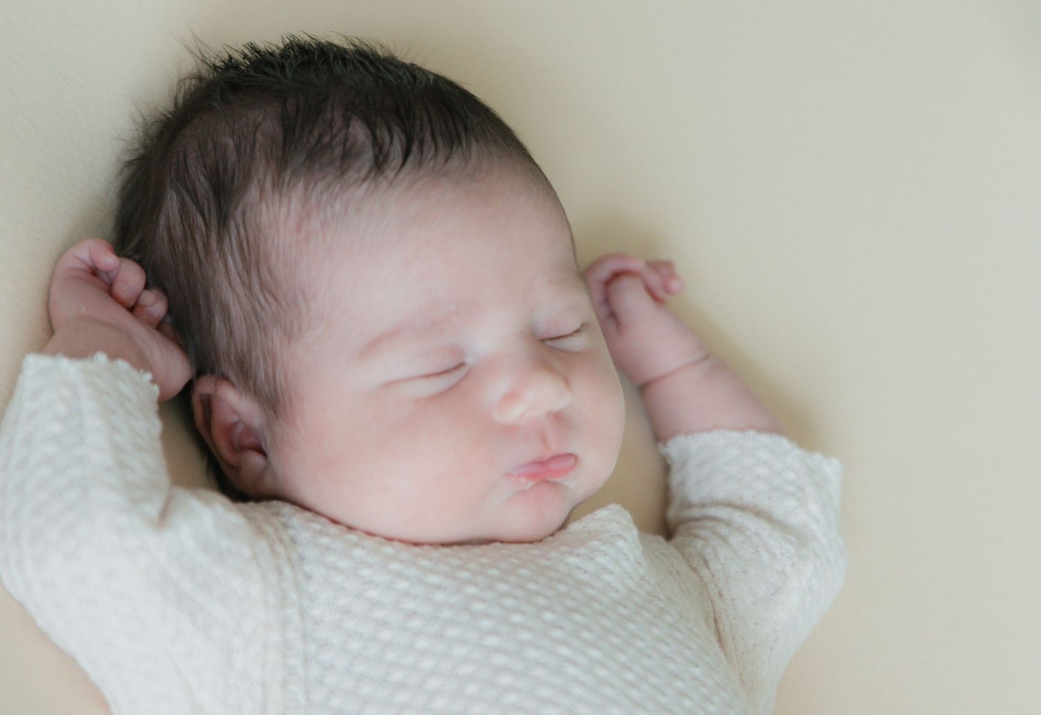 Peaceful newborn baby sleeping on a soft yellow blanket wearing a white onesie with tiny hands raised.