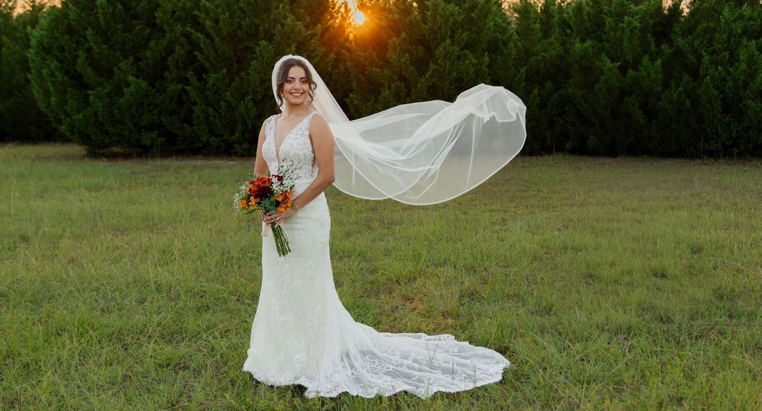 A bride in a flowing white wedding dress poses outdoors during golden hour sunset in a grassy field.