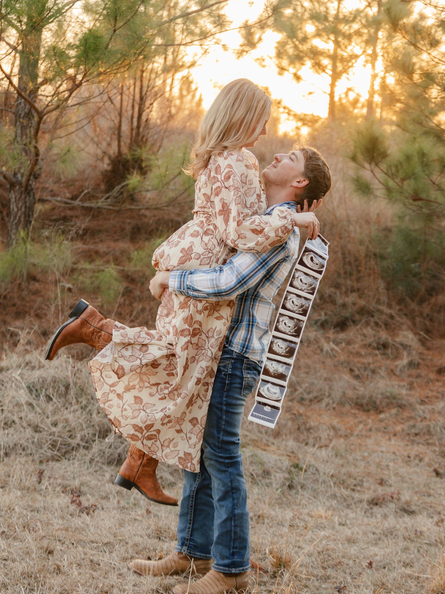 A couple in denim and floral dress dance together in a sunlit field at golden hour while their dog watches nearby.