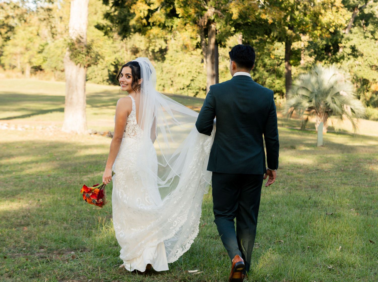 Bride in a lace gown with a flowing veil and groom in a dark suit walking through a sunny park.