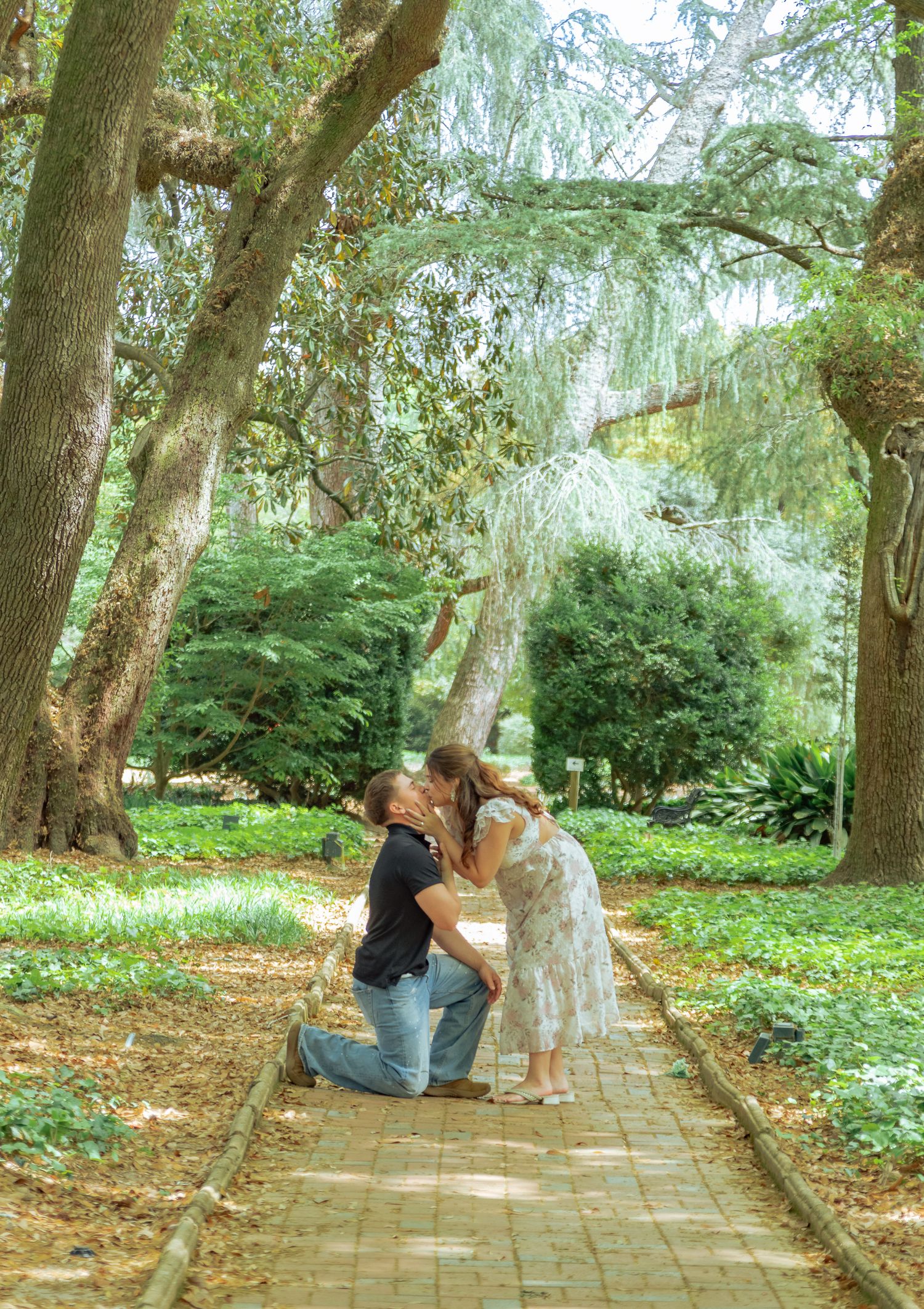Couple sharing a romantic kiss on a brick garden path surrounded by lush trees during an outdoor engagement proposal.