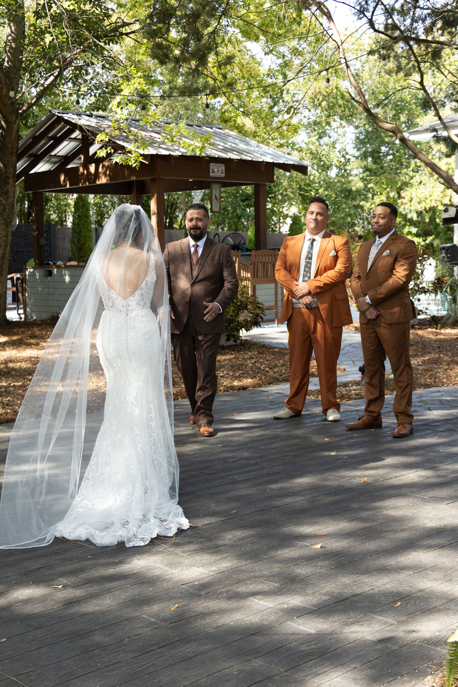 Wedding party in brown suits and flowing white dress pose for photos on a tree-lined outdoor path.
