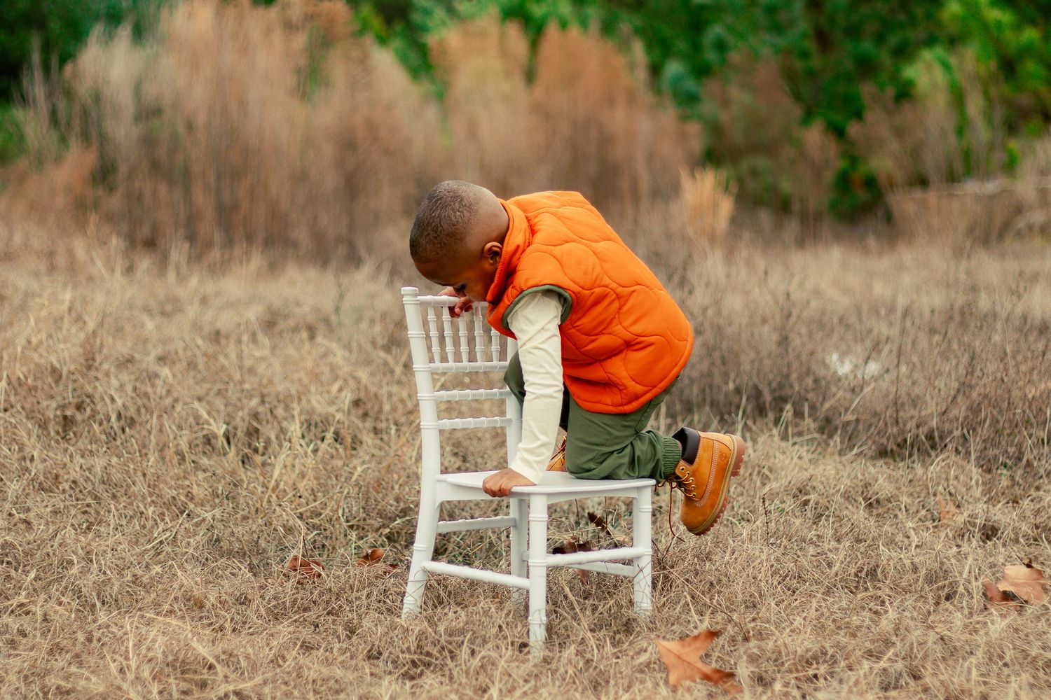 A small child in orange clothing explores a white wooden chair in a grassy field on a sunny day.