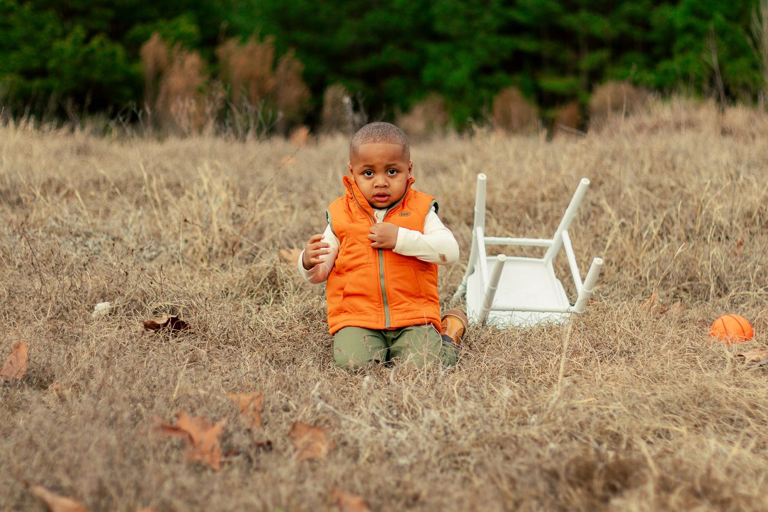 A child in an orange shirt plays in a brown grassy field with a white chair.