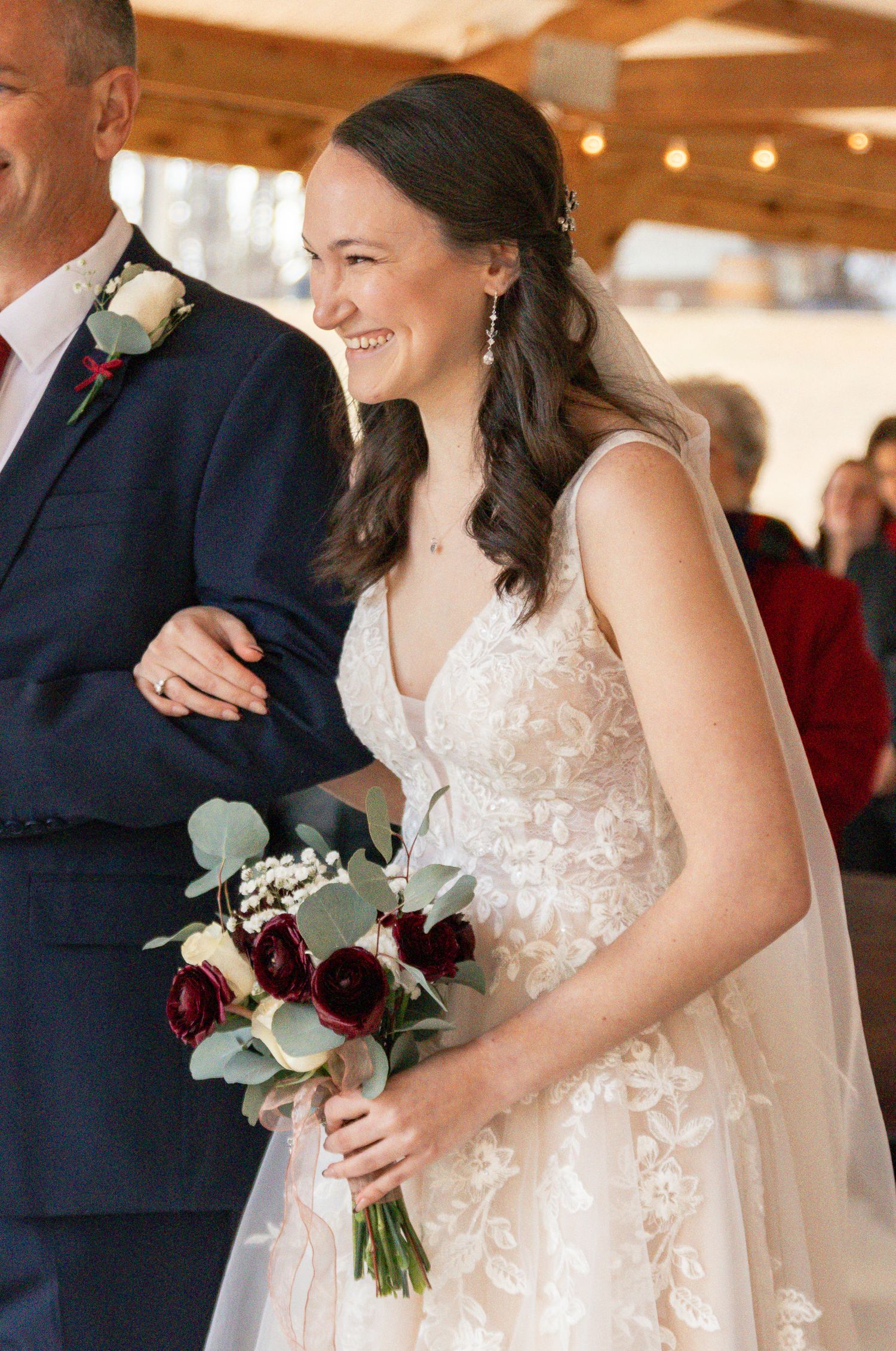 Wedding party in formal attire with white lace dress and burgundy floral bouquet at indoor venue.