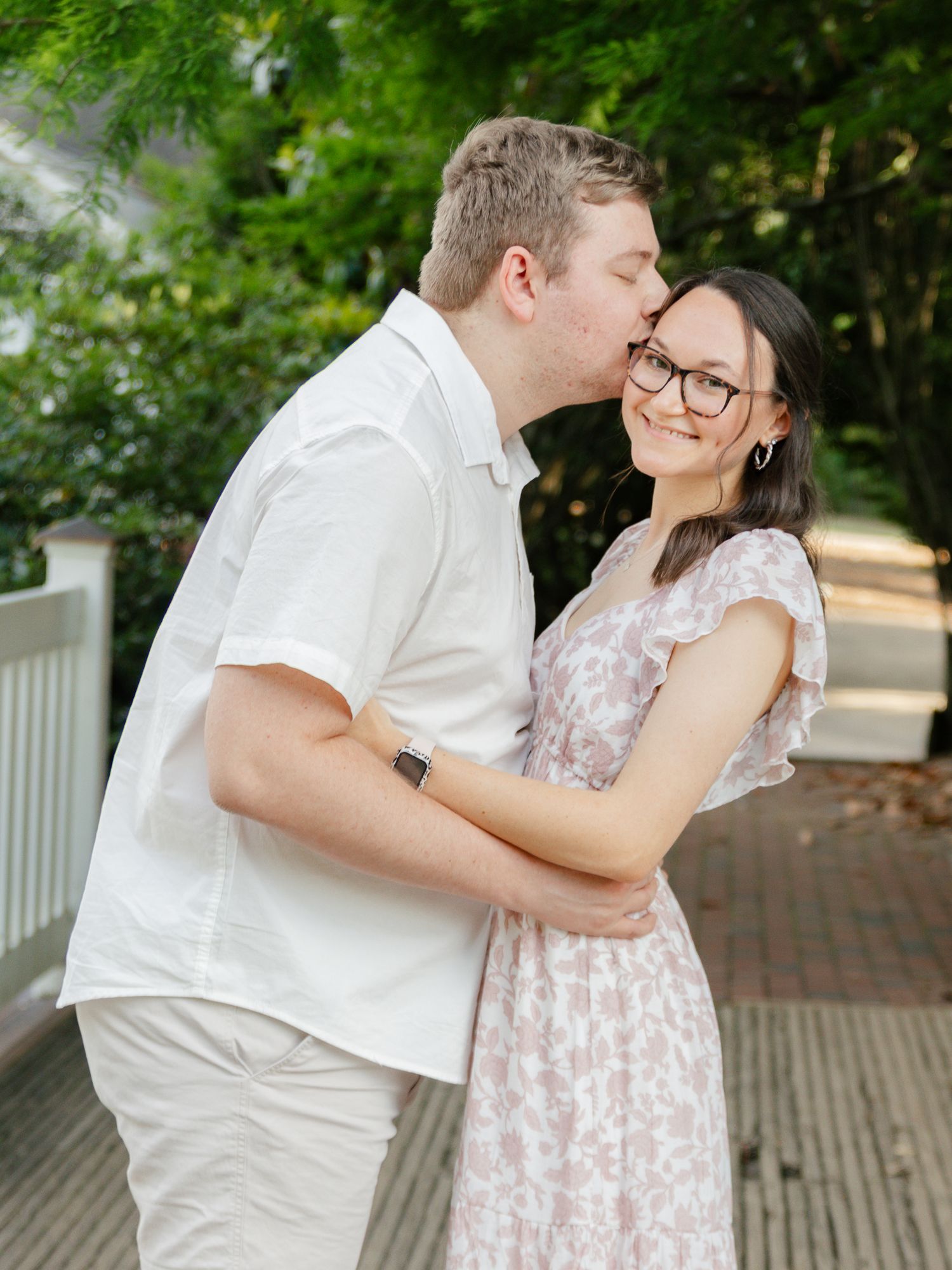 Man kisses woman on cheek as she smiles warmly during outdoor engagement session on bridge.