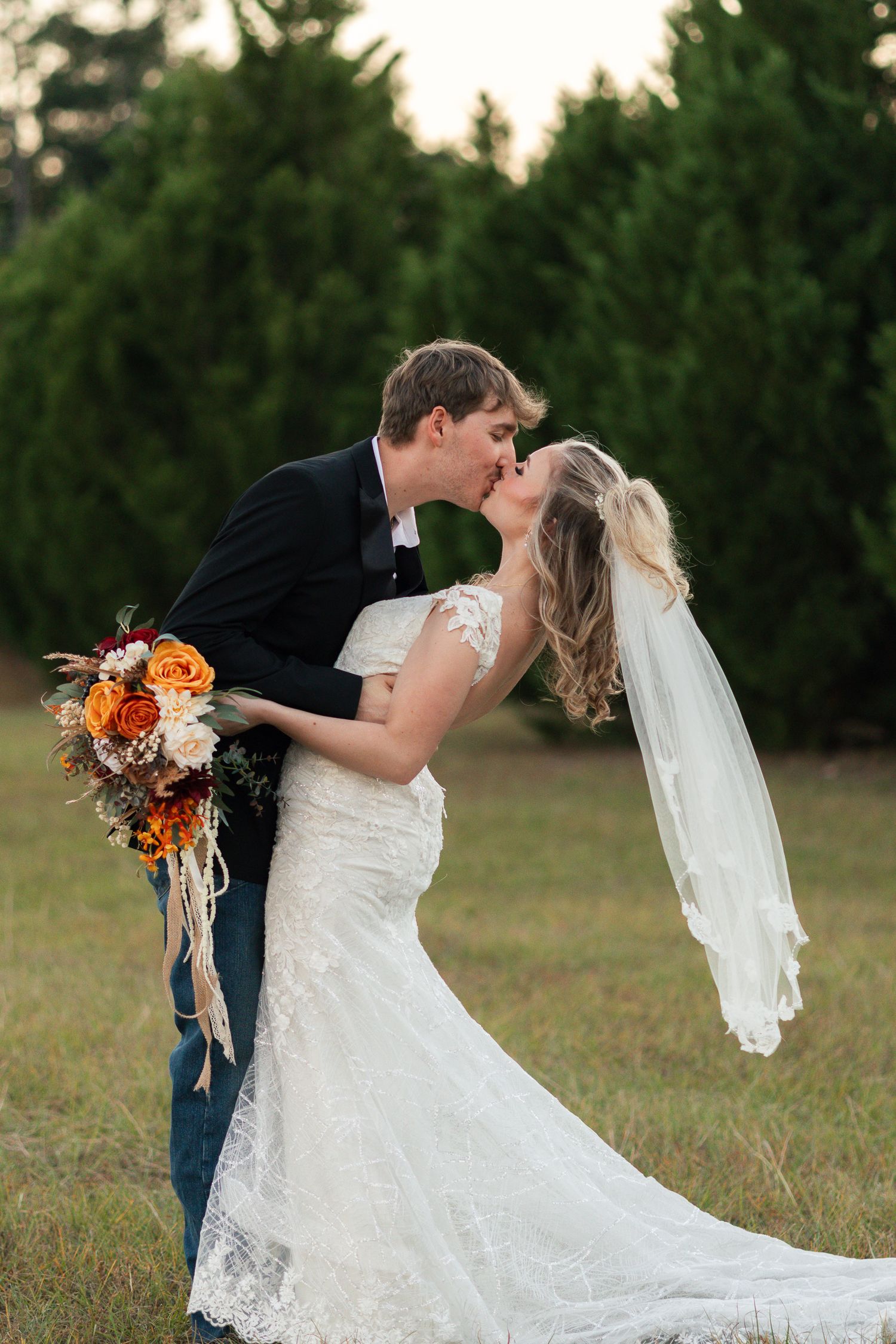 Bride and groom sharing a romantic dip kiss outdoors holding an orange floral bouquet at sunset.