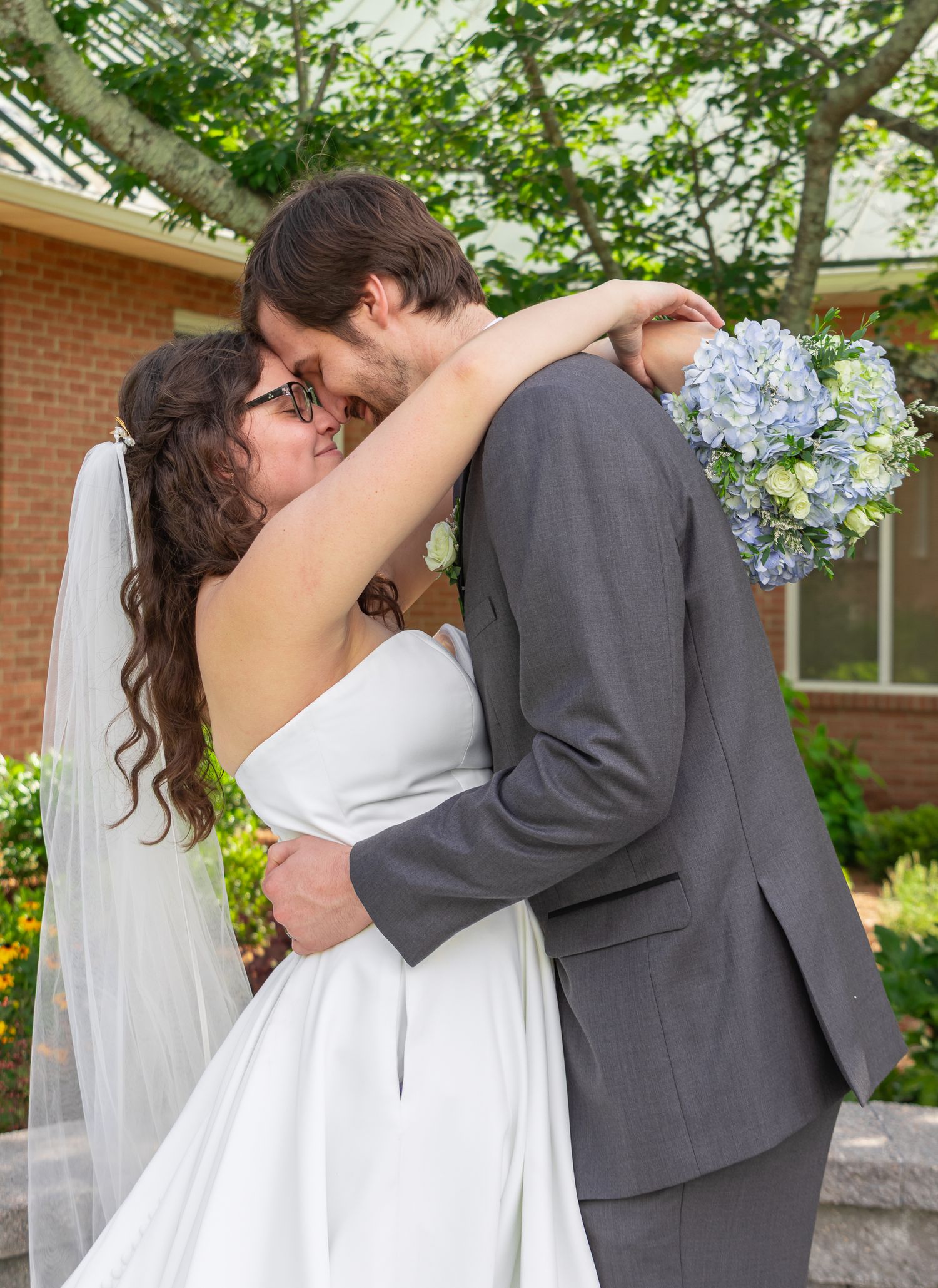 Bride and groom sharing a romantic kiss outdoors on their wedding day, with the bride holding a blue hydrangea bouquet.