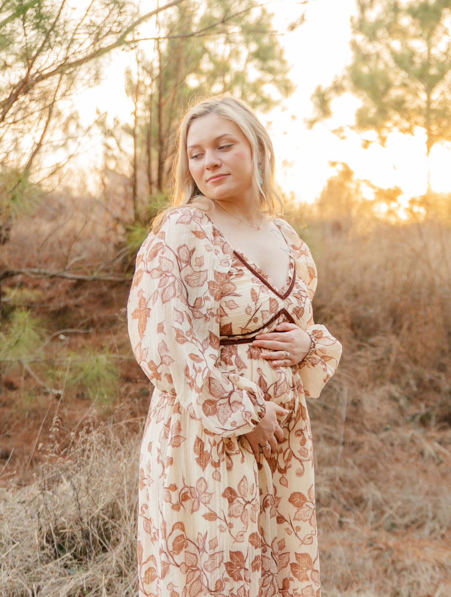 A woman in a floral dress poses outdoors during golden hour sunset with soft lighting and natural scenery.