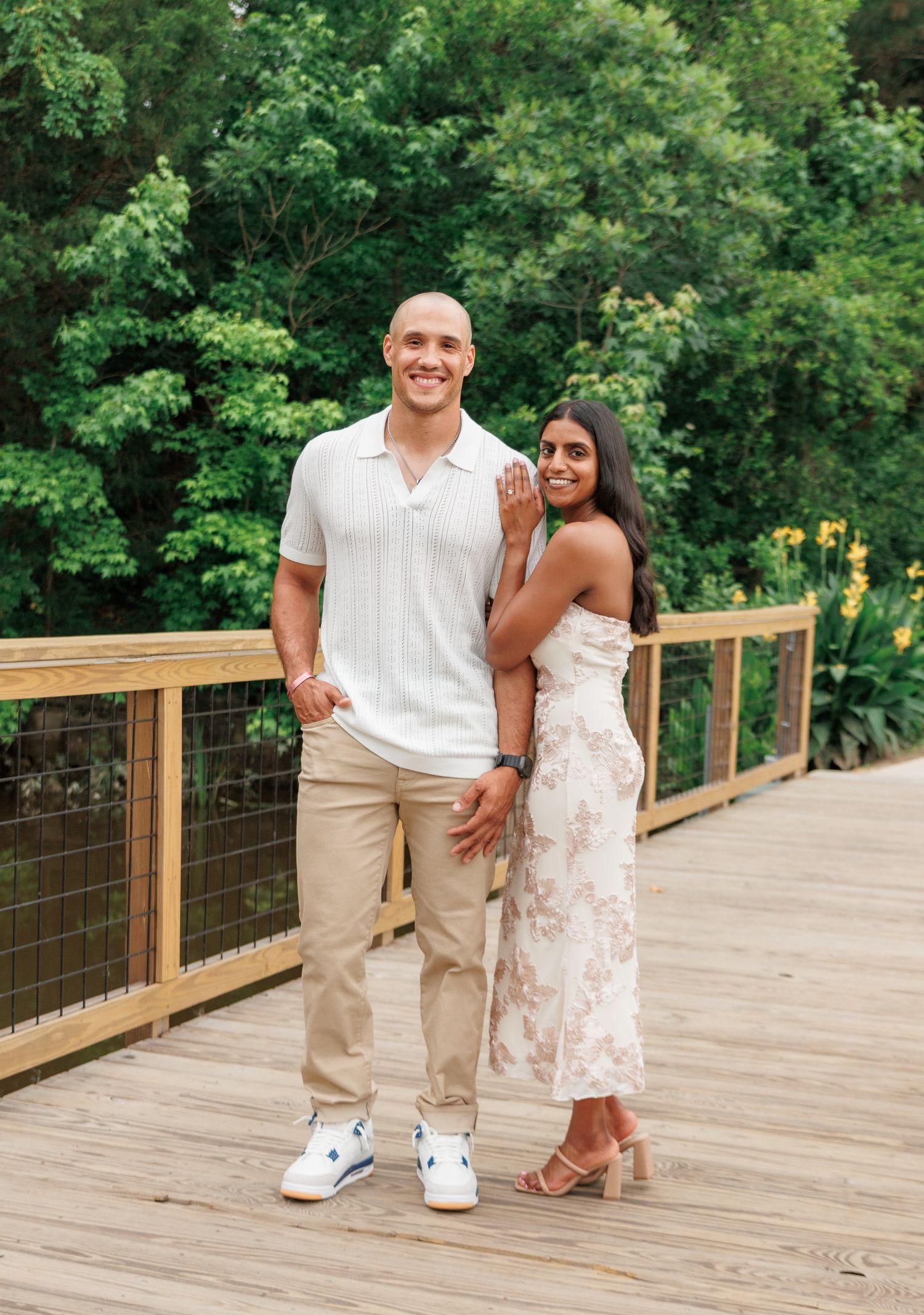 Smiling couple posing together on a wooden bridge with green trees in the background.