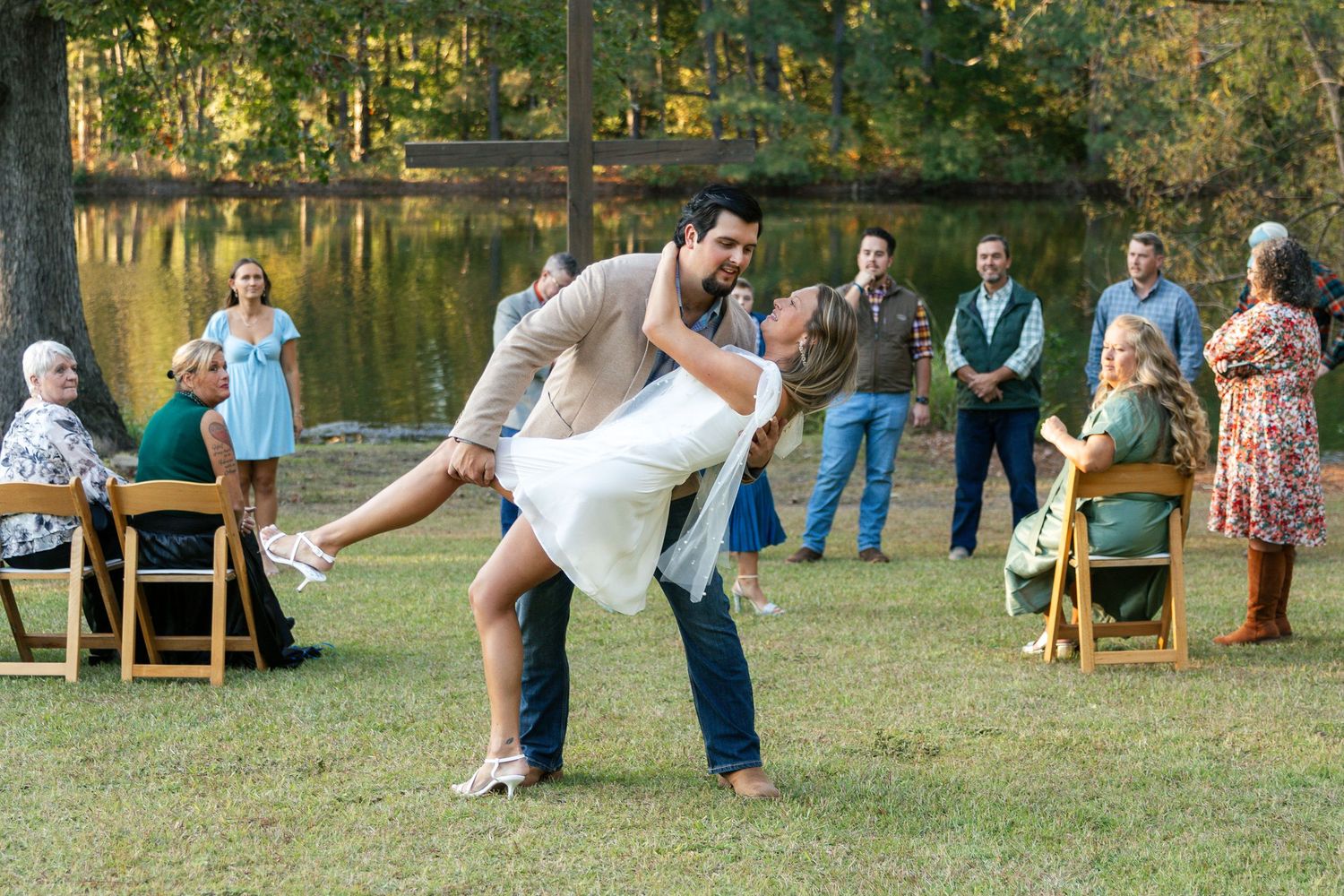 Outdoor wedding ceremony in a wooded setting with wooden chairs arranged on grass and guests gathered around.
