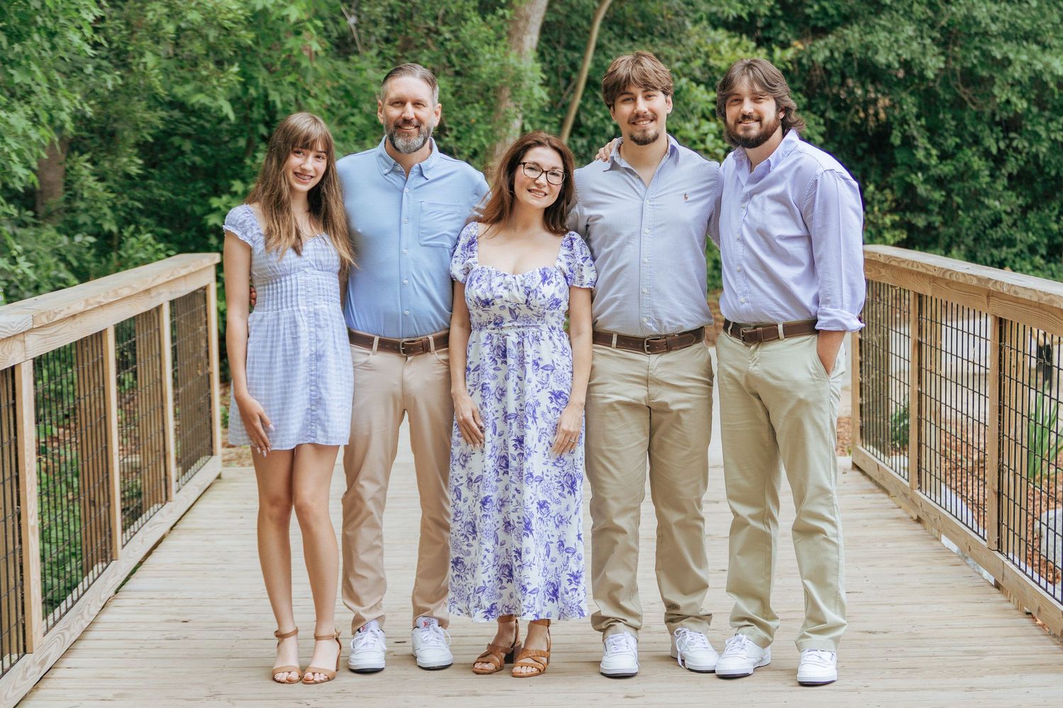Family of five smiling together on a wooden bridge surrounded by lush green trees.