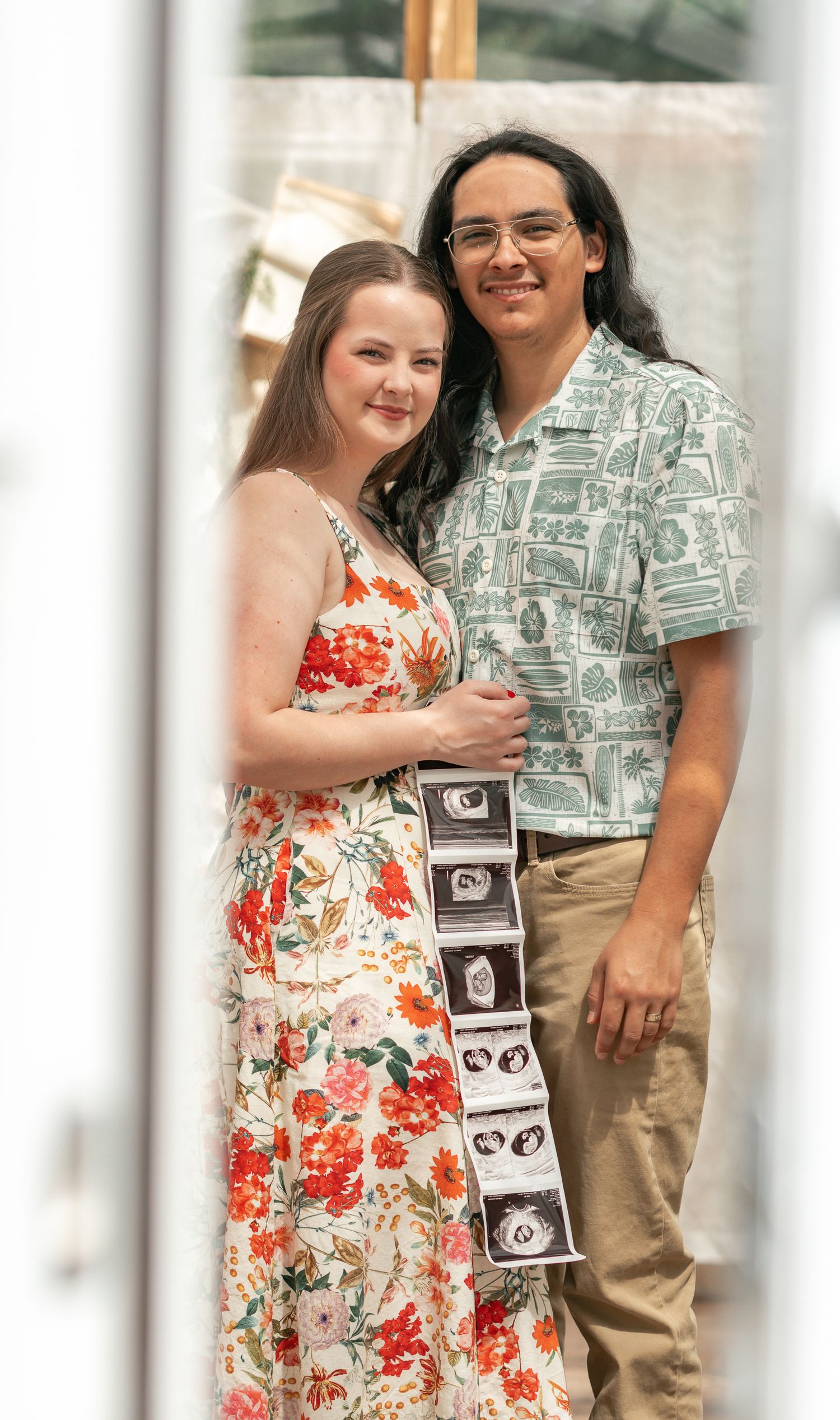 Smiling couple holding ultrasound photos at a pregnancy announcement photoshoot, woman in floral dress, man in Hawaiian shirt.