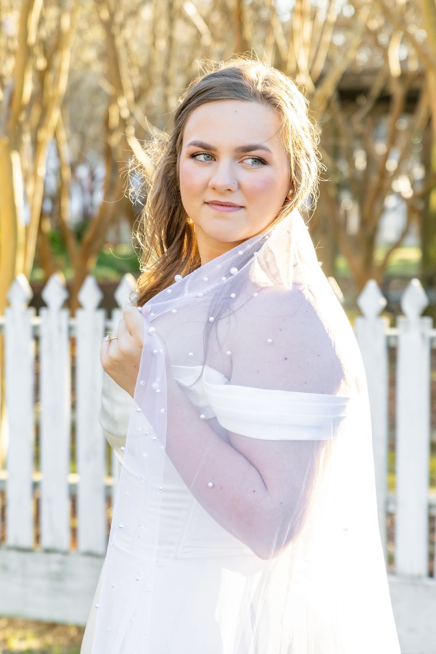 Bride in off-shoulder white gown with pearl-dotted veil posing near white picket fence outdoors.