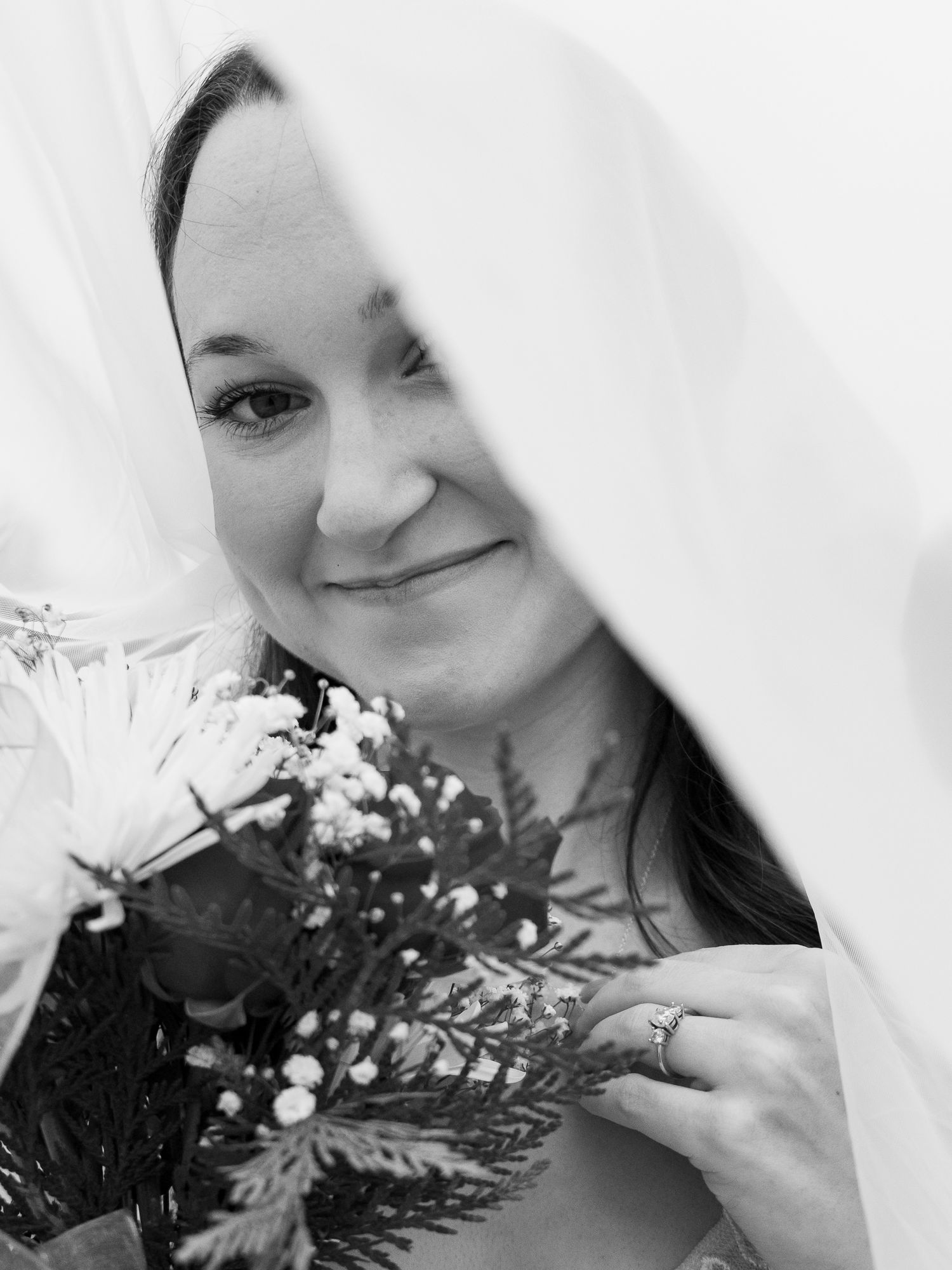 Black and white portrait of a smiling bride holding a floral bouquet beneath her veil.