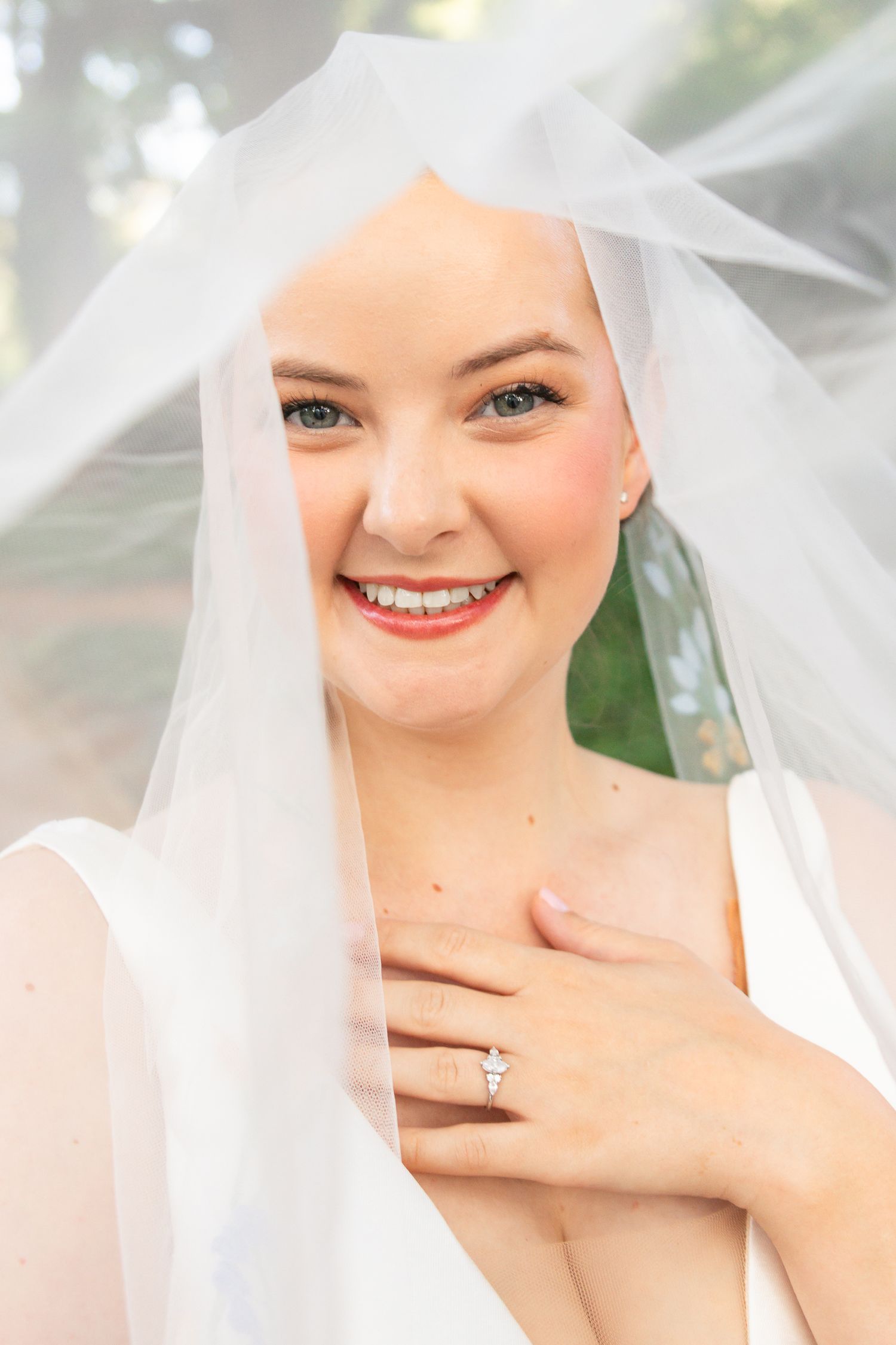 Beautiful bride smiling under a white veil, showcasing her diamond engagement ring.