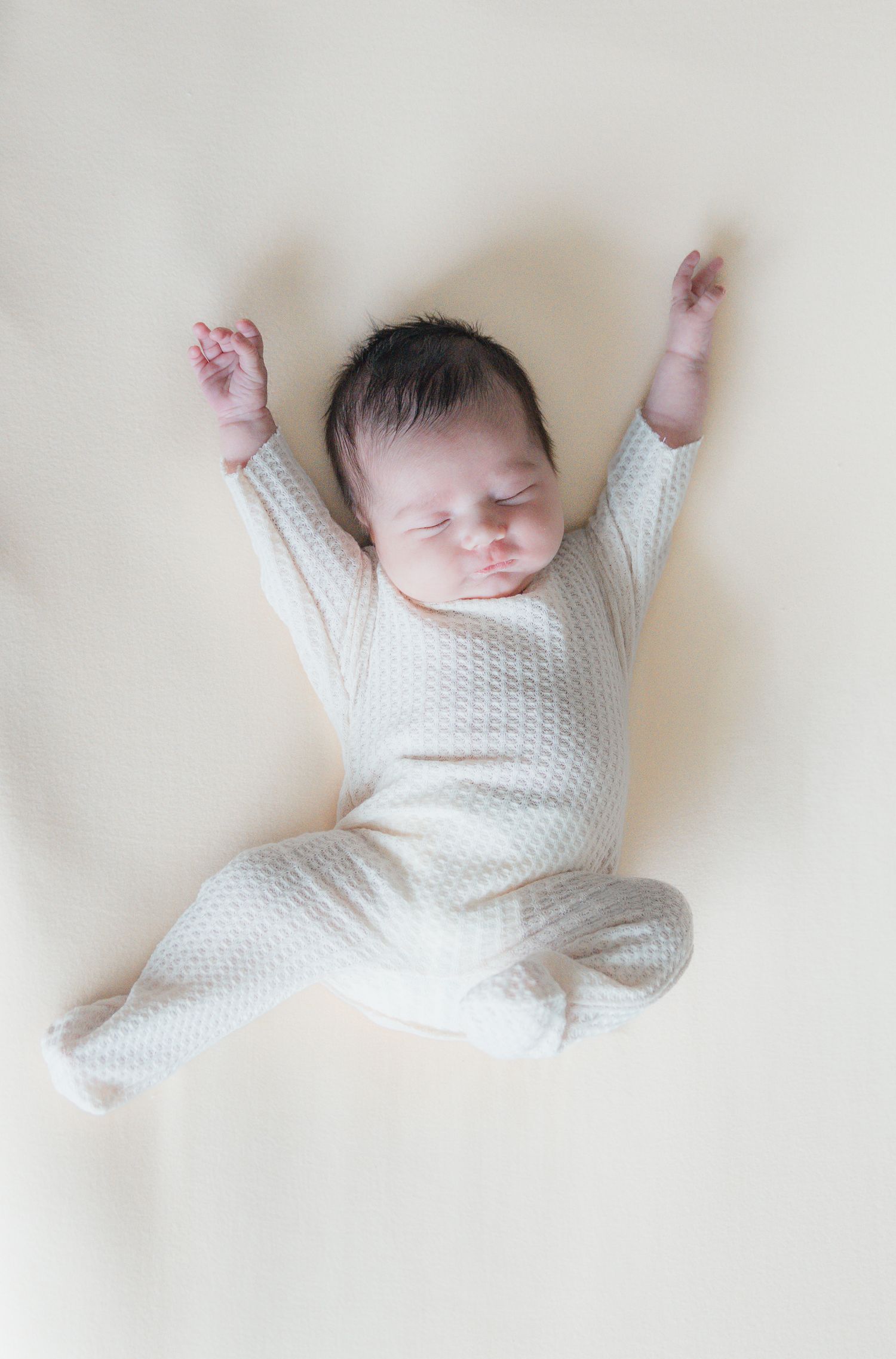 Adorable newborn baby in white knit onesie sleeping with arms stretched out on a soft cream surface.