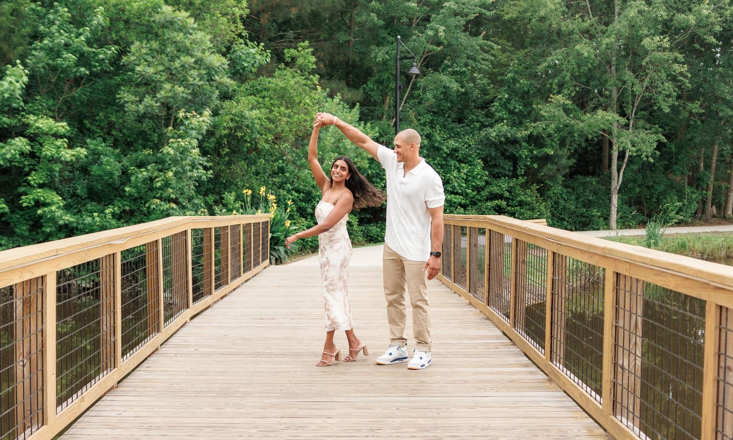 Couple dancing with raised arms on a wooden bridge surrounded by lush green trees.