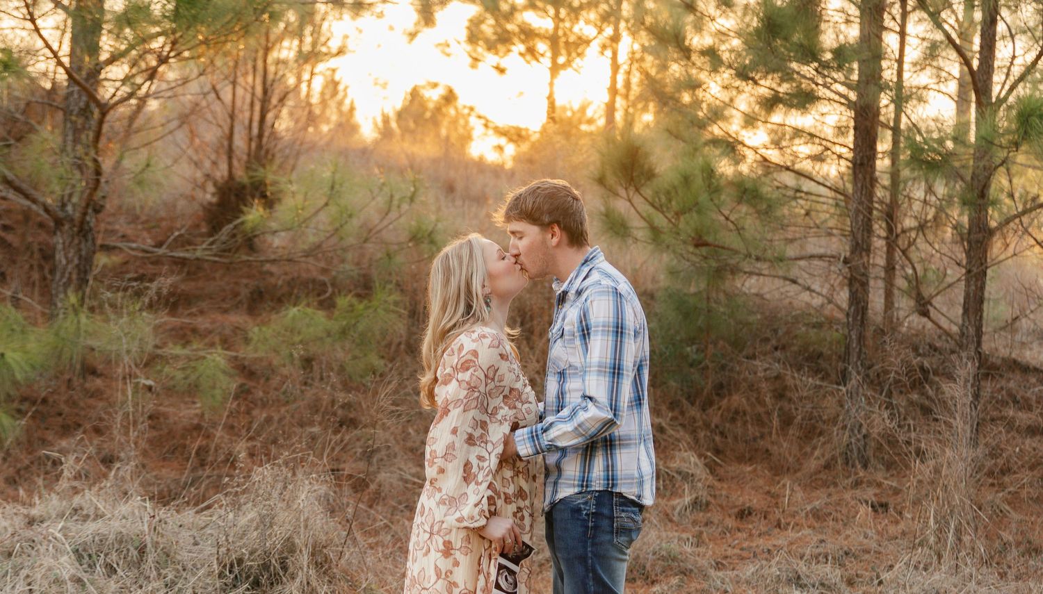 A romantic sunset moment captured between a couple in a floral dress and plaid shirt among pine trees.