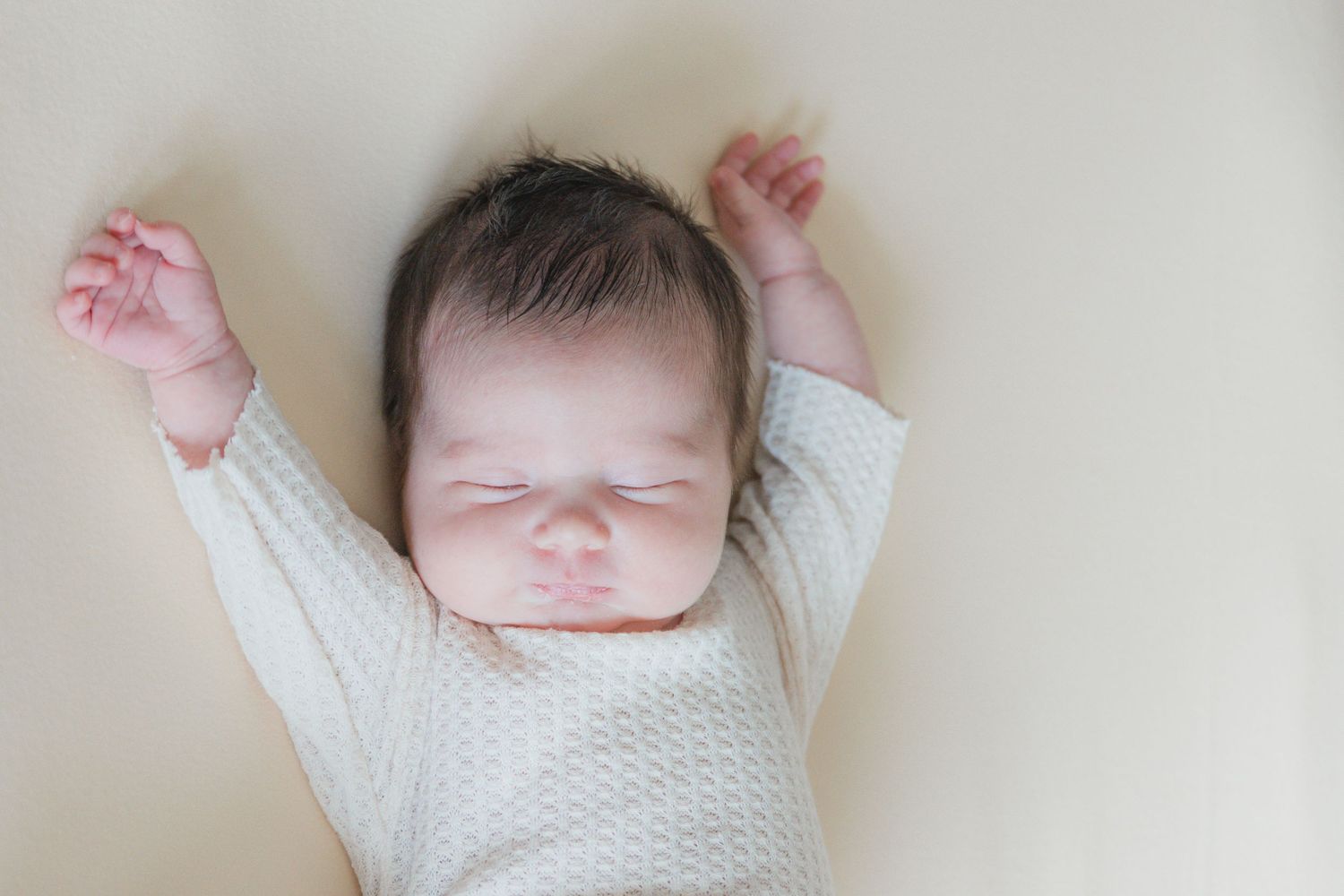 Serene newborn baby with dark hair sleeping with arms raised in a cozy white knit onesie.