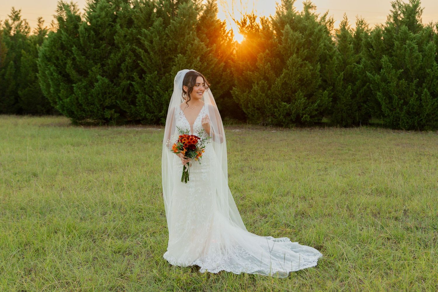 A bride in a flowing white lace gown holds a vibrant red bouquet against a sunset backdrop in a grassy field.