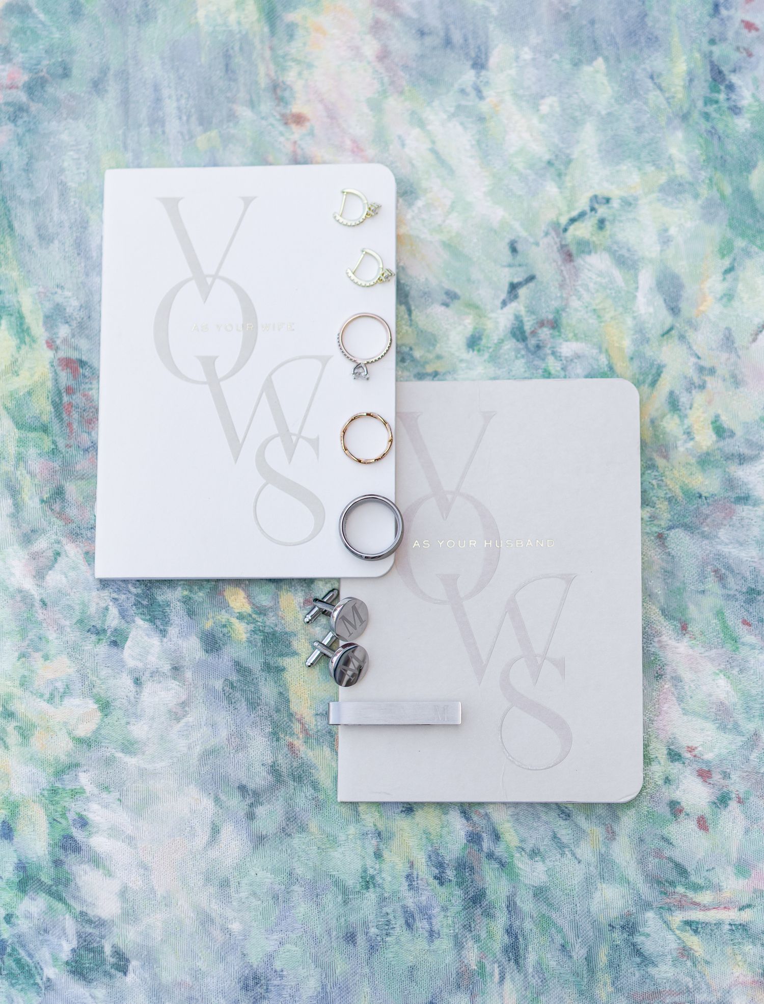 Two white vow books with monogram lettering displayed with wedding rings on a soft floral watercolor background.