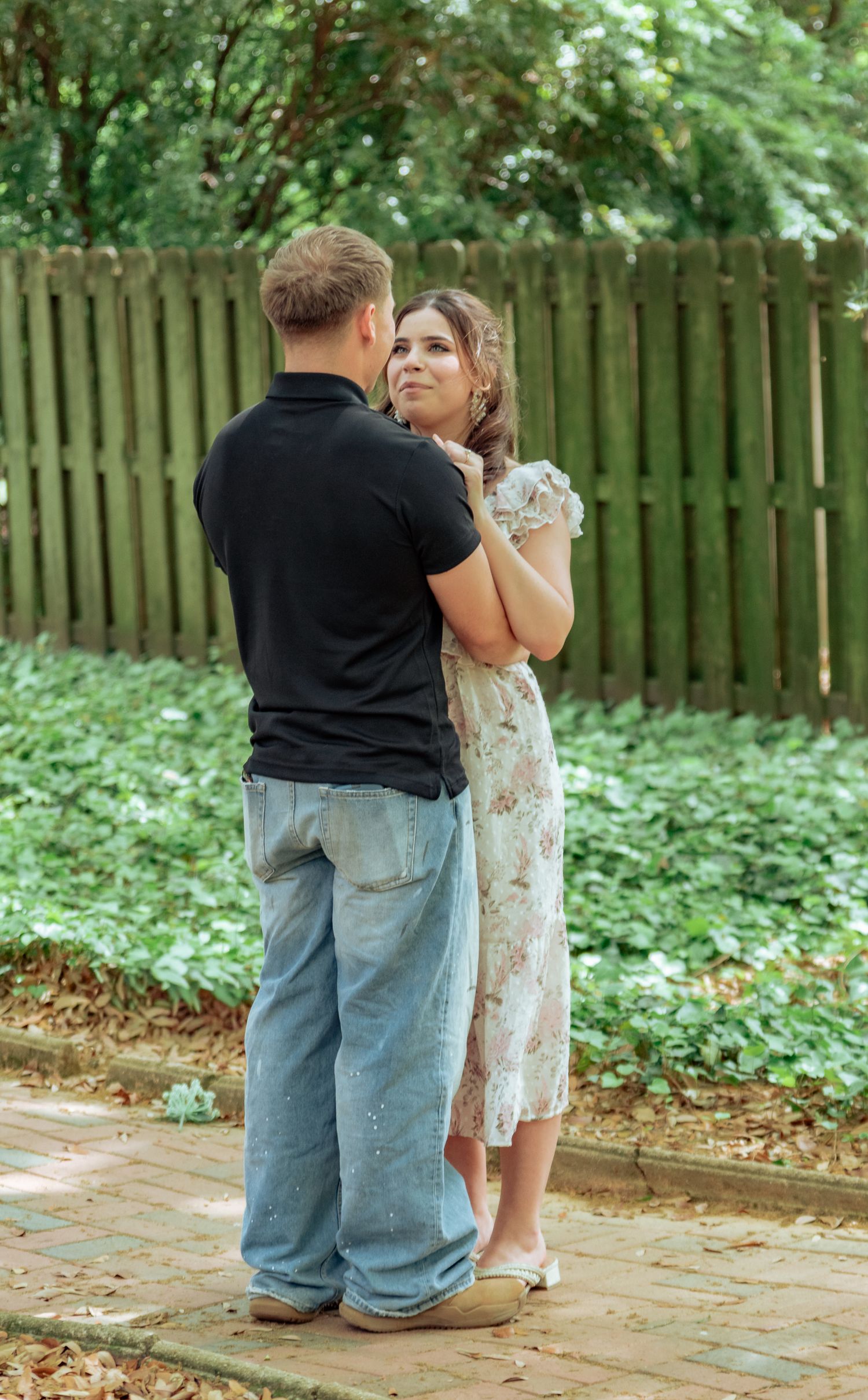 Couple standing together in a garden, man in black polo and jeans, woman in floral dress, near a wooden fence.