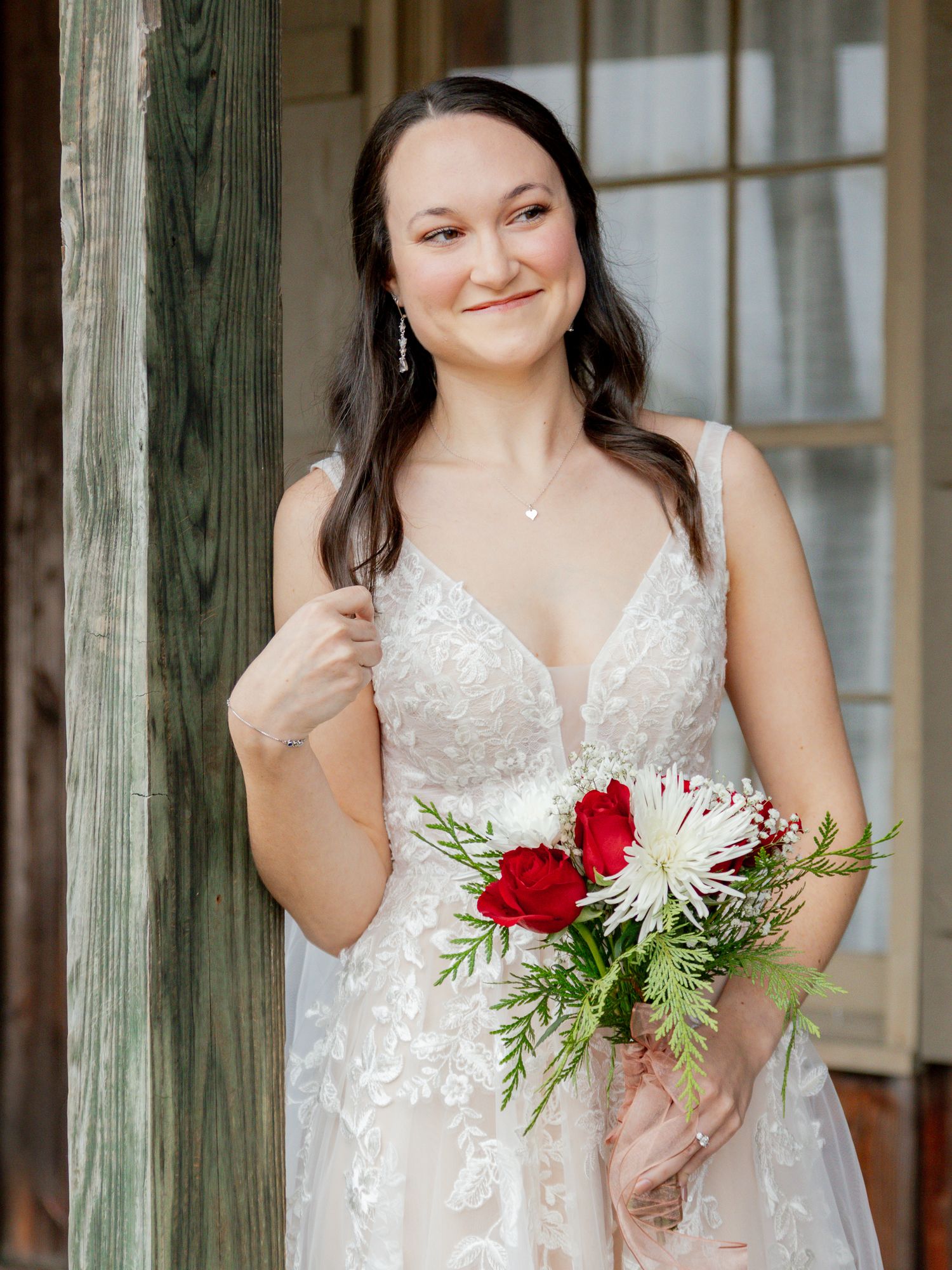 A smiling bride in a lace wedding gown holds a bouquet of red roses while posing near a rustic wooden post.