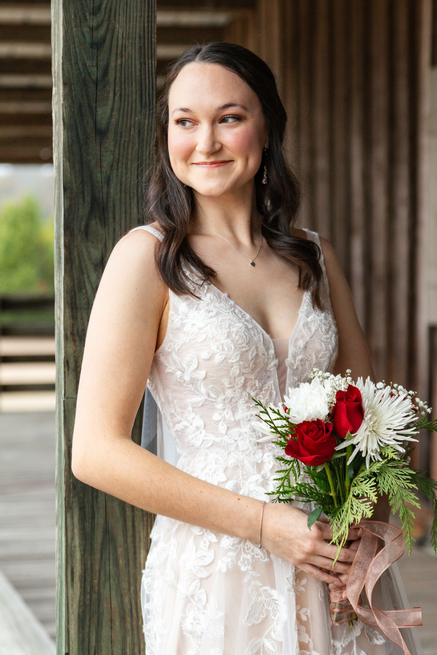 Smiling bride in lace wedding dress holding red roses and white flowers on a wooden porch.