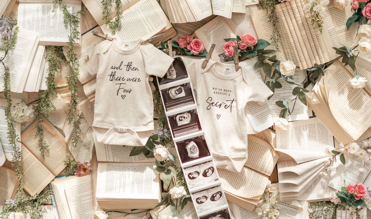 Two baby onesies and ultrasound photos displayed among open books and floral decorations for a book-themed baby shower.
