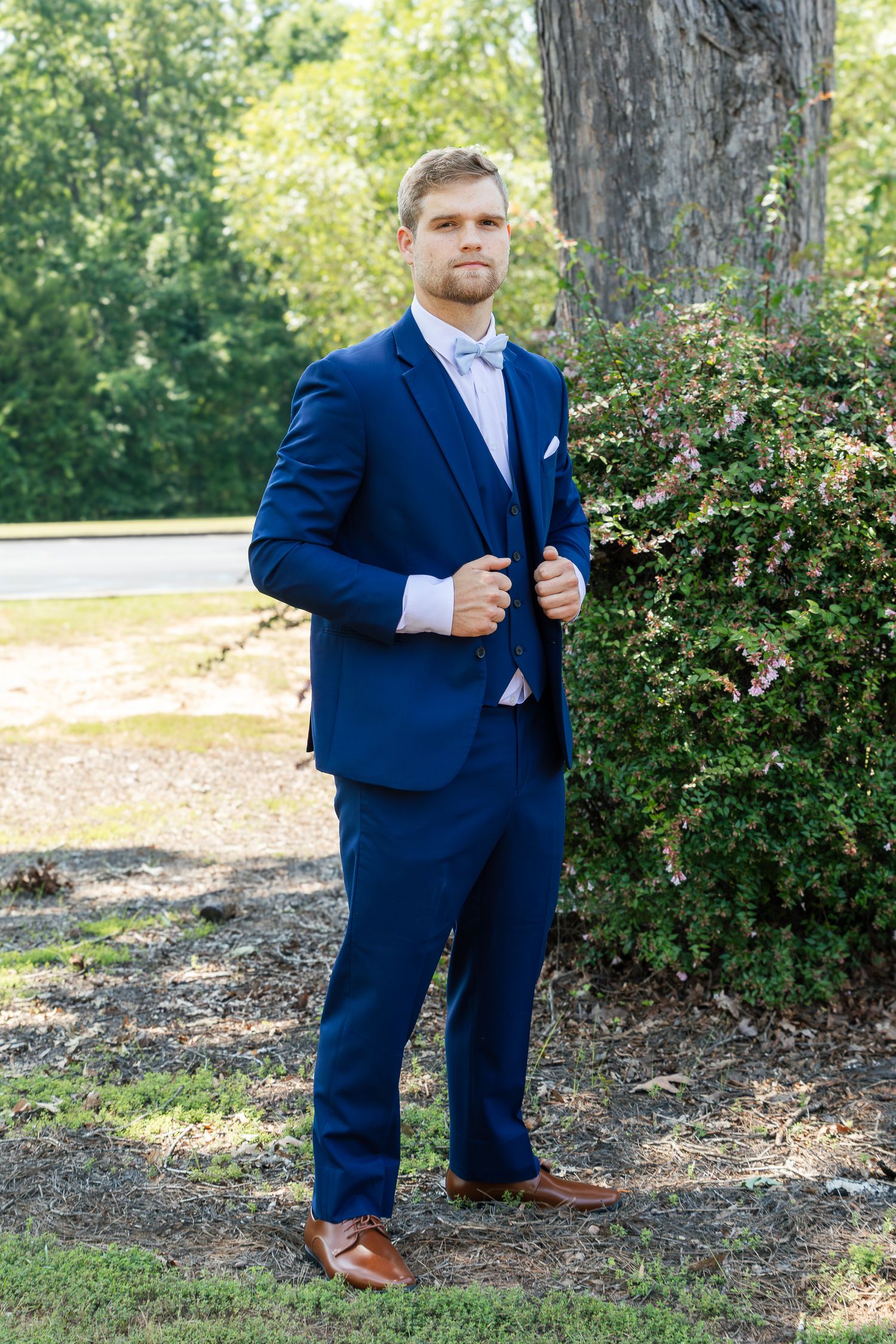 A person in a navy blue suit and brown shoes poses in front of green bushes and trees in an outdoor setting.