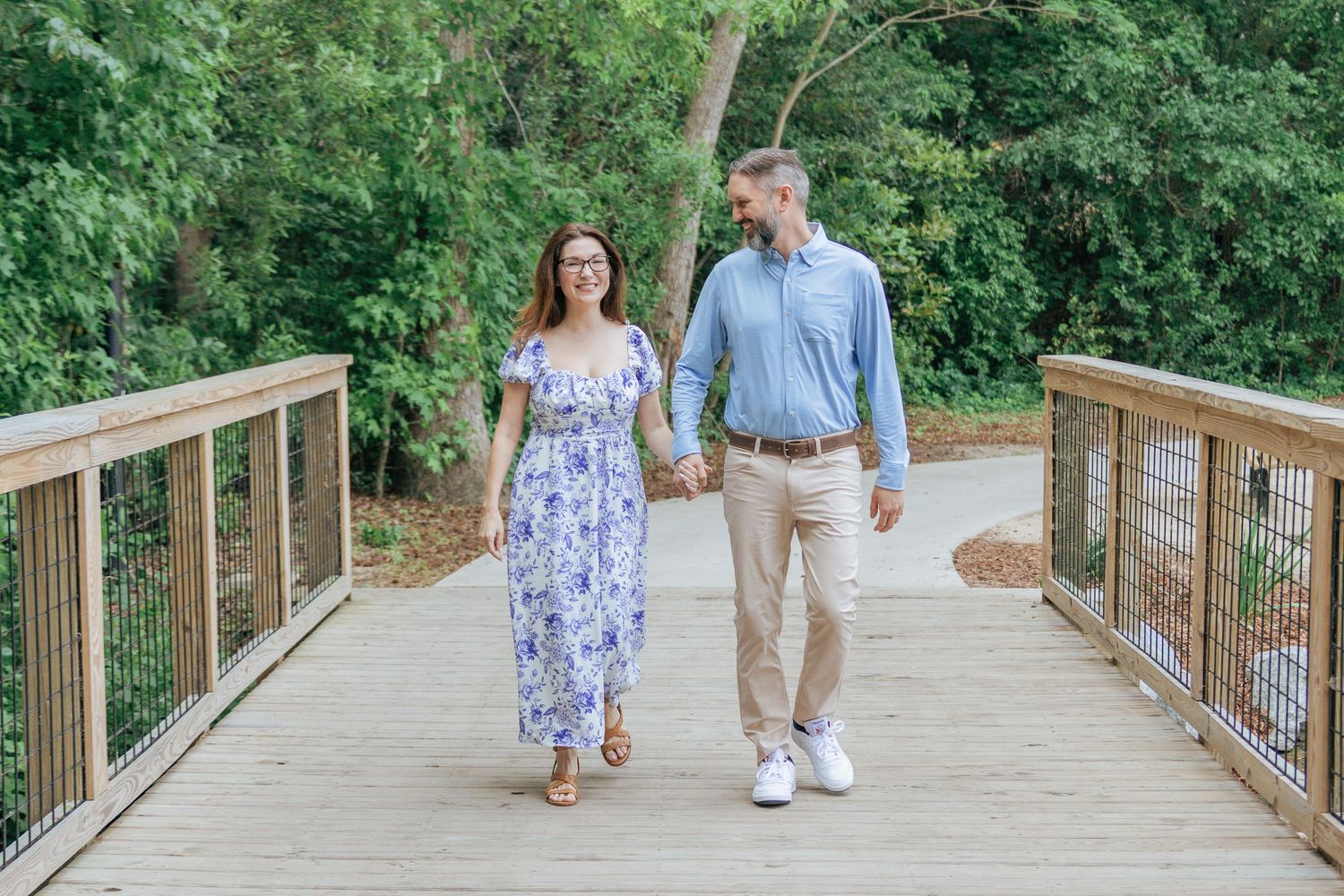 Engaged couple walks hand-in-hand across wooden bridge surrounded by lush green trees.