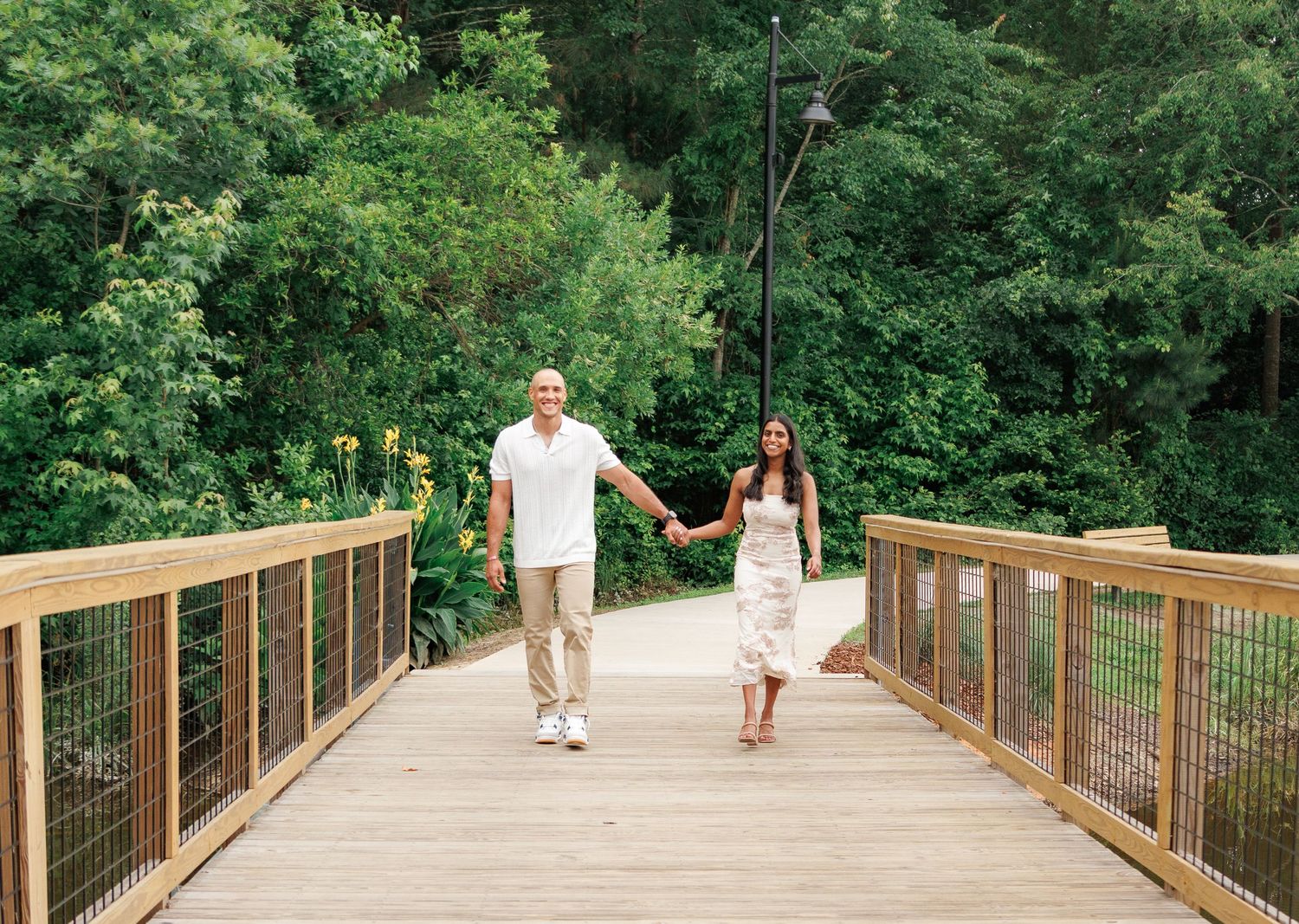 Happy couple holding hands walking on wooden bridge with green trees in background.