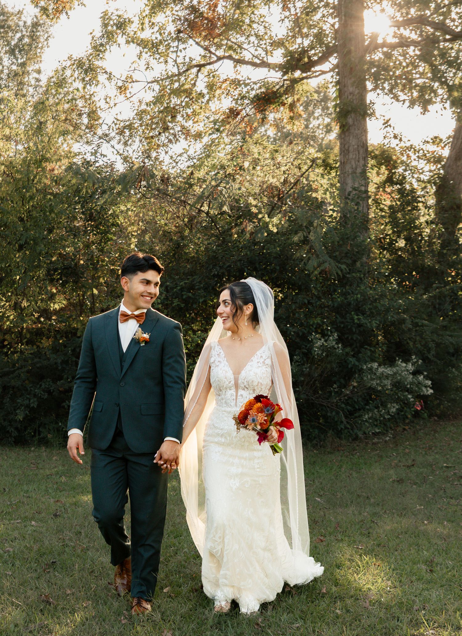 A couple in wedding attire hold hands while walking outdoors during golden hour with autumn trees in the background. Lexington Wedding