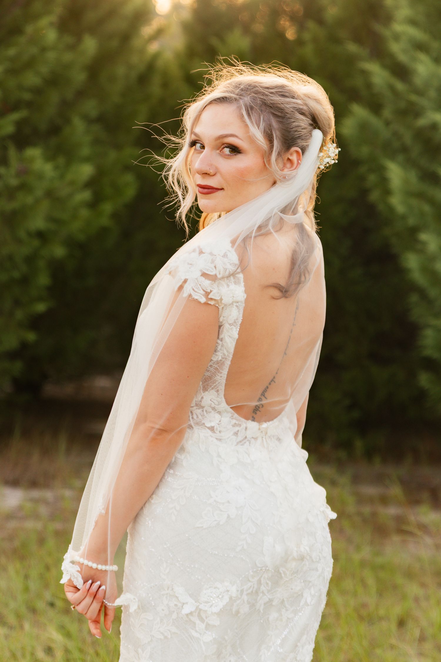 A bride in a white lace wedding dress poses outdoors during golden hour with sunlight filtering through trees.