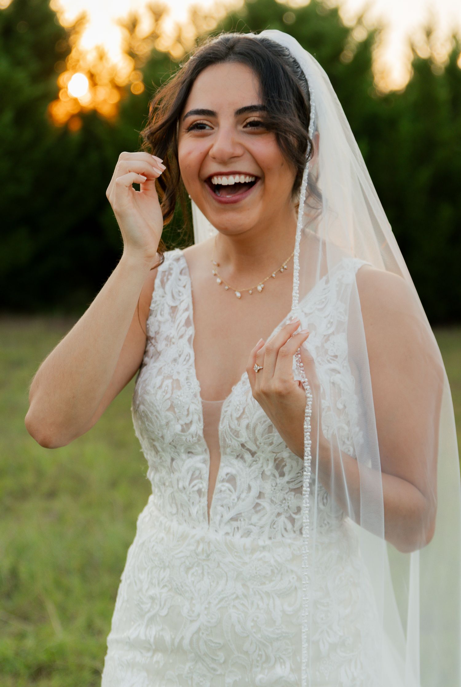 Joyful bride in lace gown laughing while holding her veil at golden hour in a field.