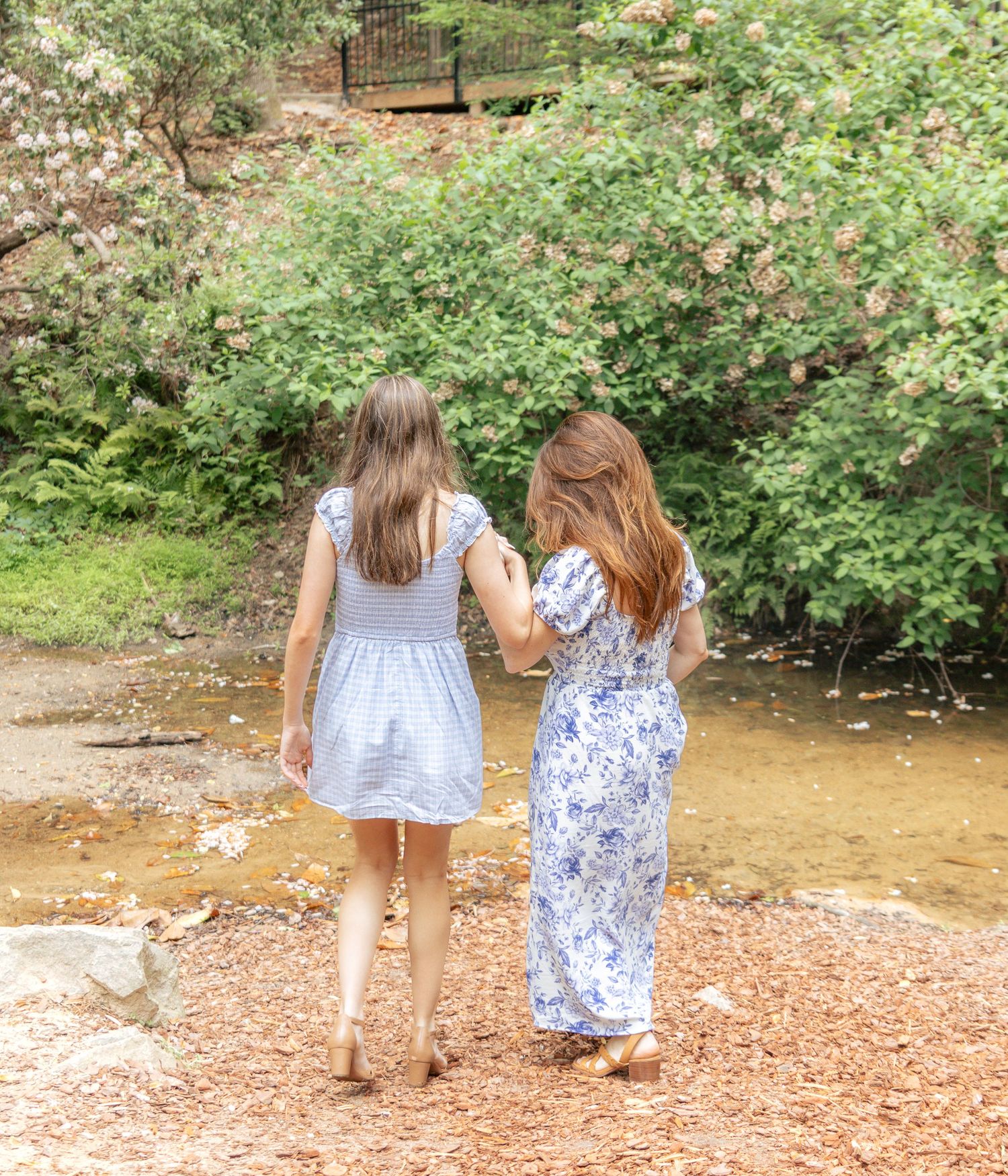 Two girls in summer dresses walk hand-in-hand toward a peaceful creek surrounded by blooming trees.
