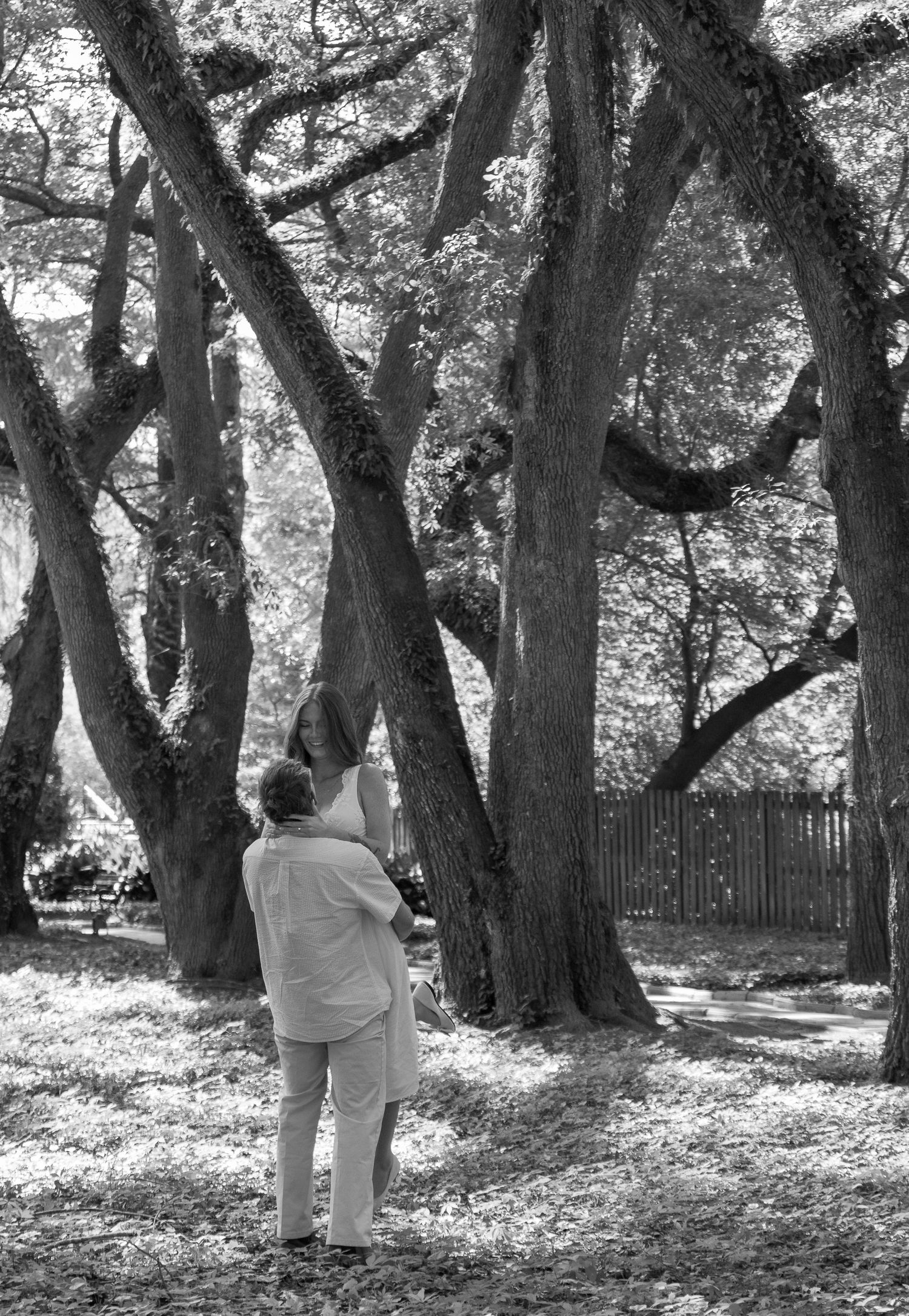Black and white engagement portrait of a couple embracing beneath large trees at Hopeland Gardens in Aiken, South Carolina.