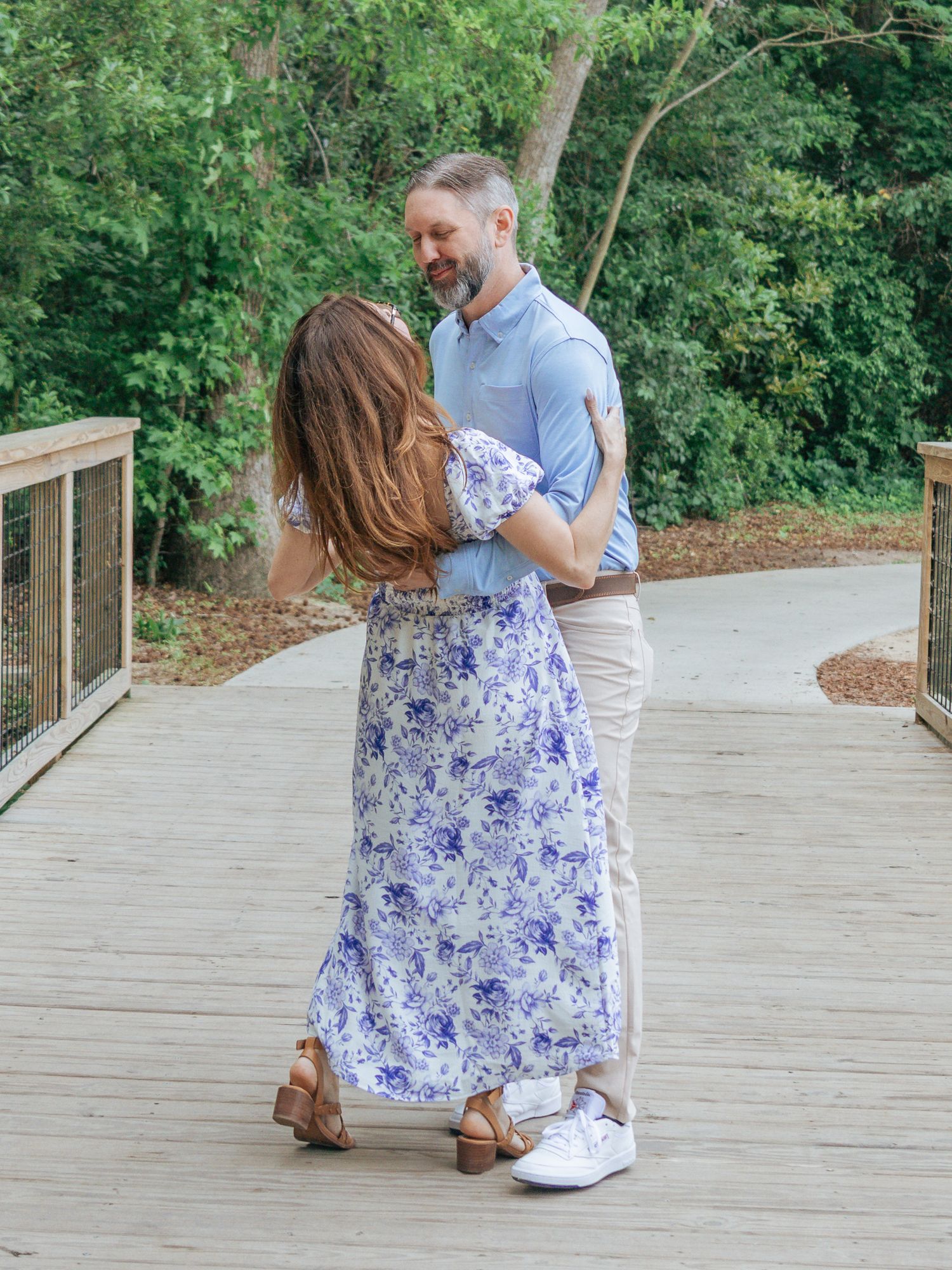 Couple dancing on a wooden bridge surrounded by lush greenery, woman in floral maxi dress, man in blue shirt.