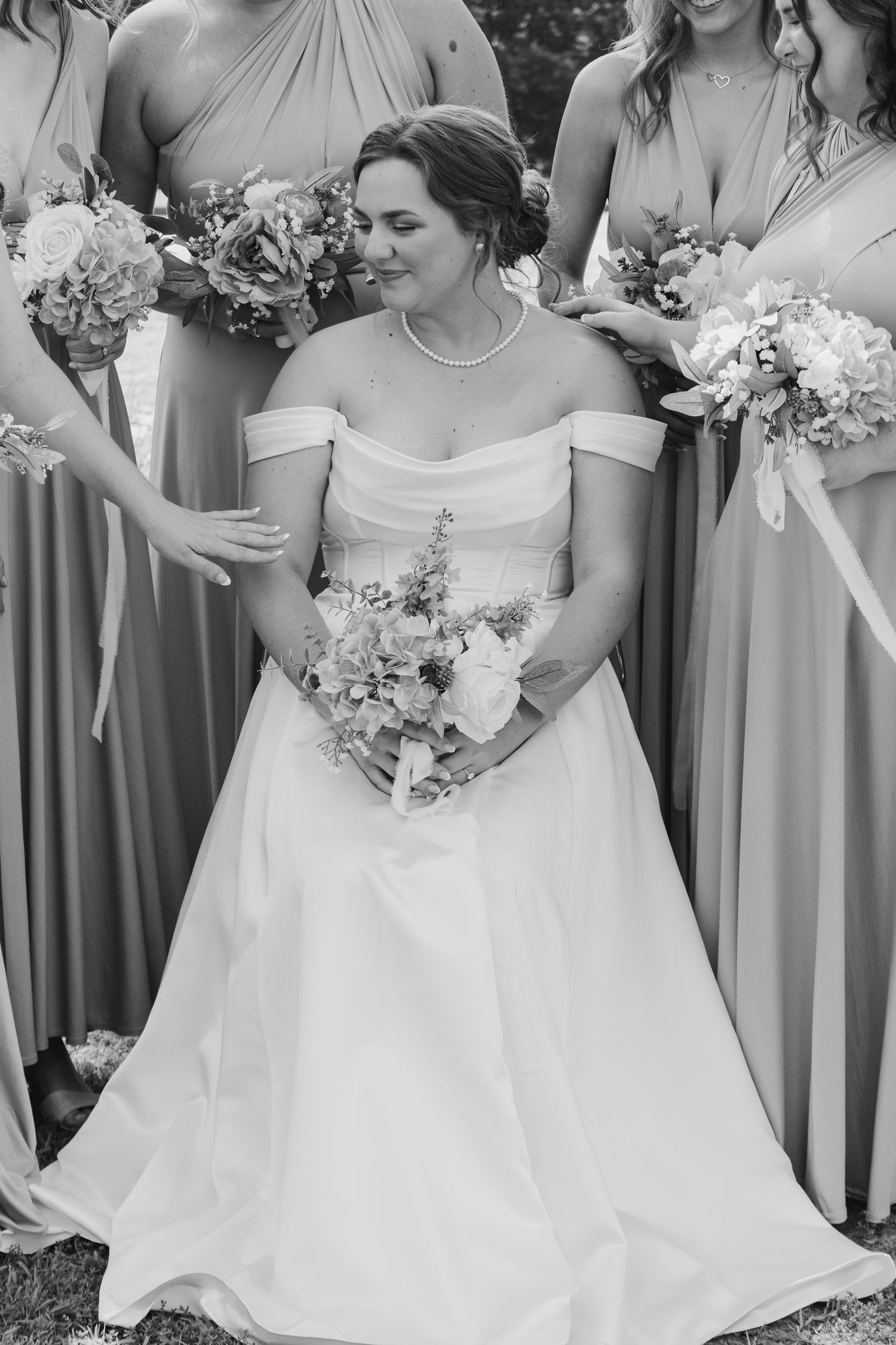 Black and white photo of a bride in an off-shoulder gown holding a bouquet surrounded by bridesmaids.