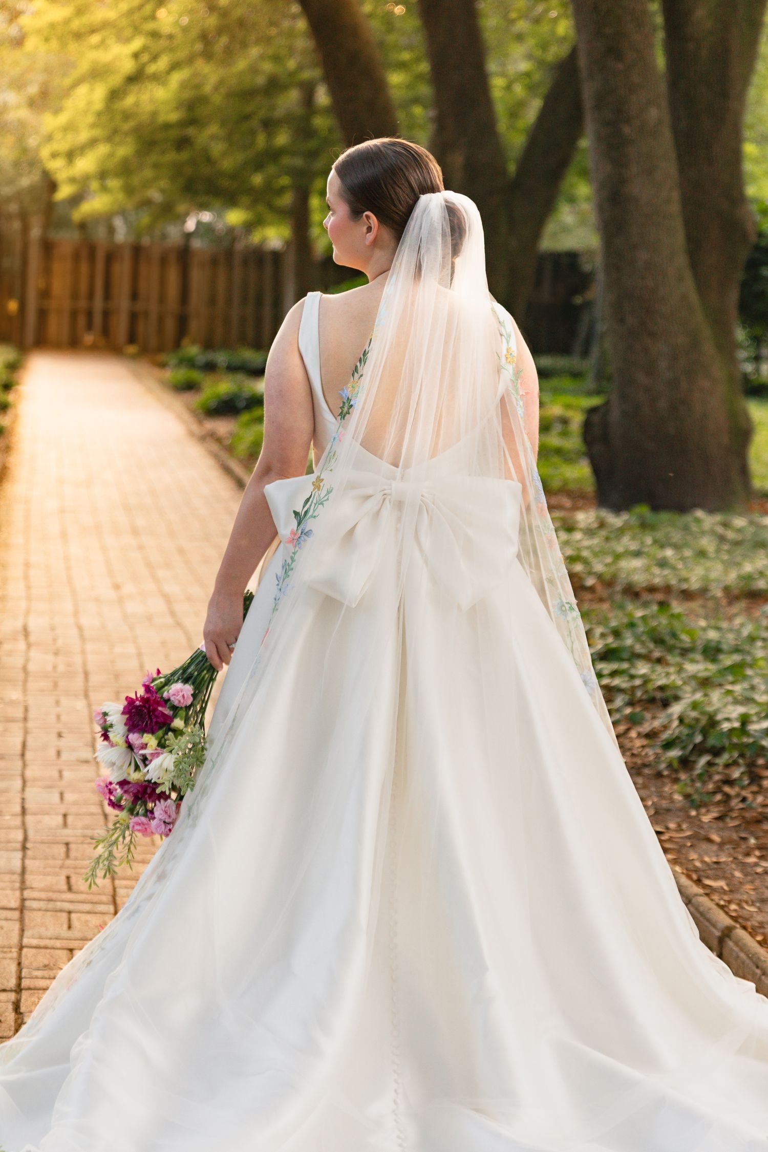A bride in a white ballgown with large bow walks down a sunlit brick path holding a colorful bouquet.