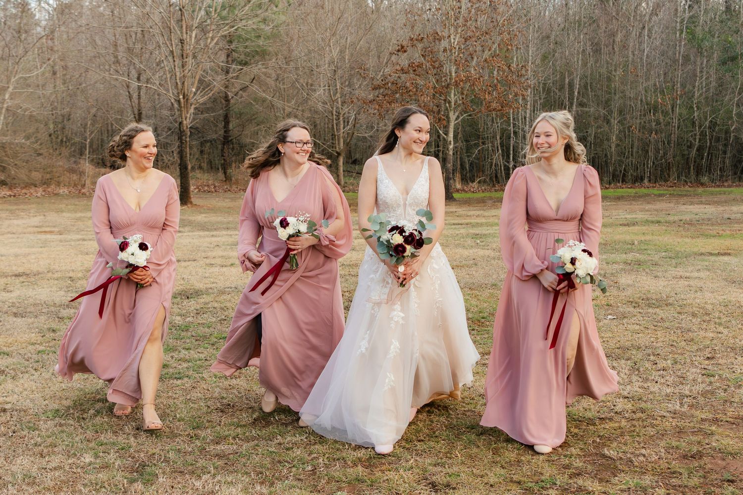 A group in dusty pink bridesmaid dresses and a white wedding gown walk together in a field during autumn.