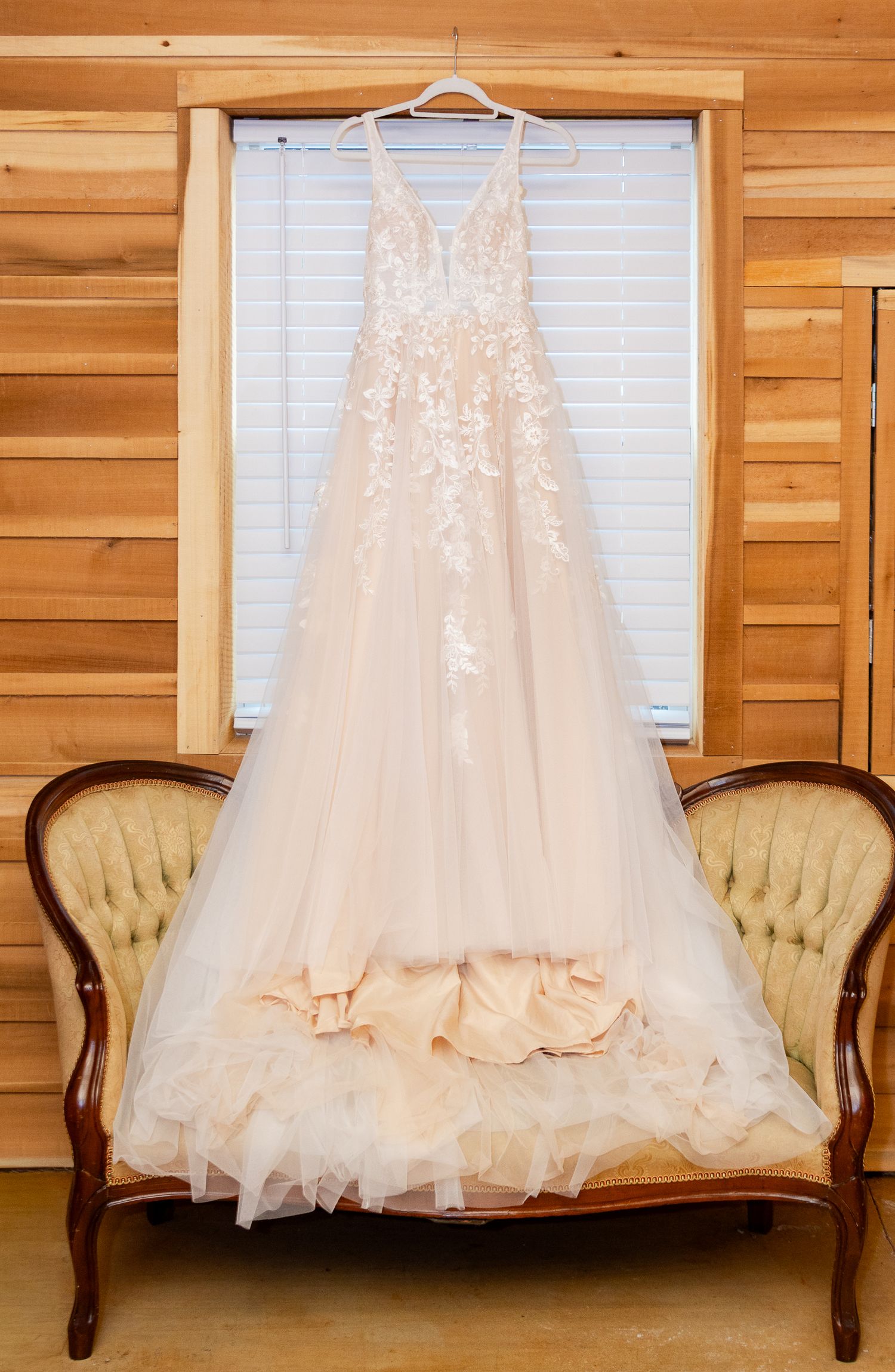 A white wedding dress with lace details hangs in front of wooden paneled walls and windows in a rustic setting.