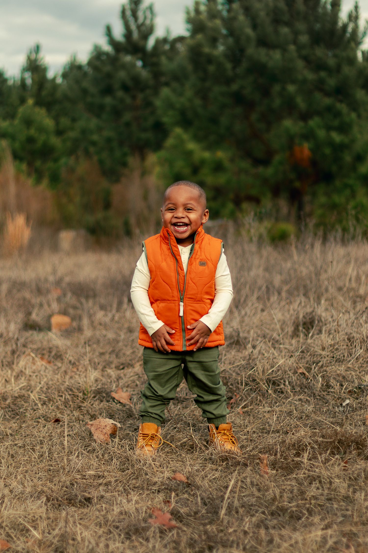 A child wearing an orange vest and boots stands in a field with pine trees in the background.