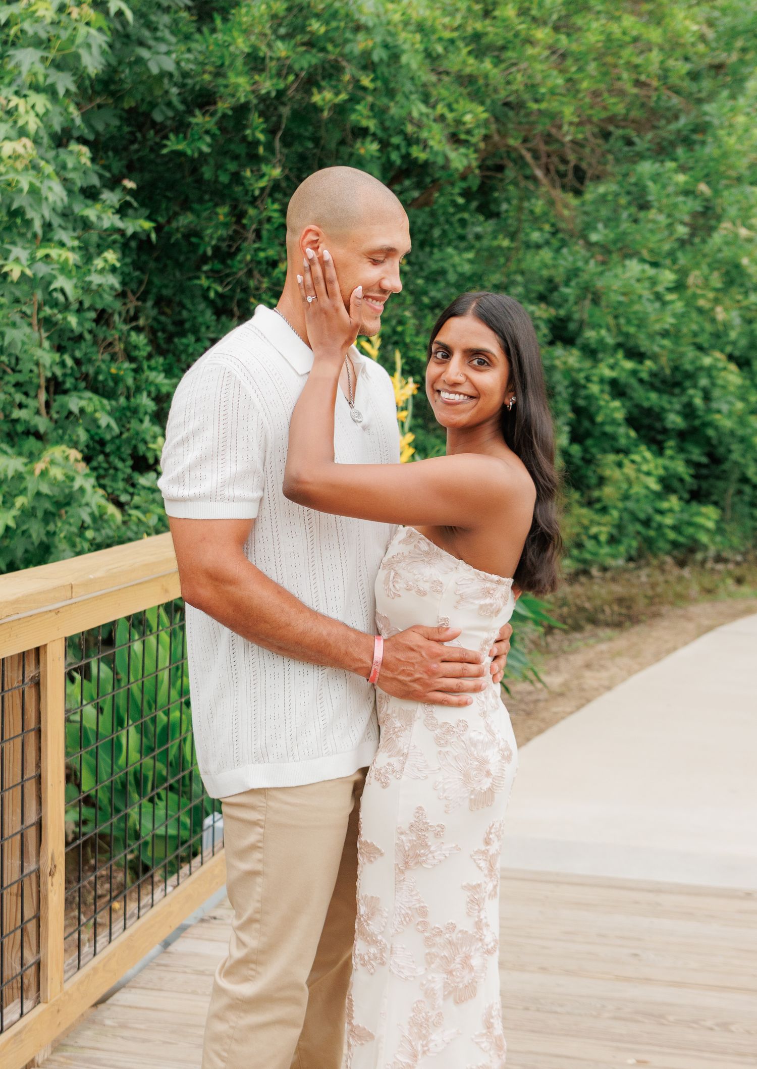 A couple embraces on a wooden bridge surrounded by lush greenery, smiling warmly in summer attire.