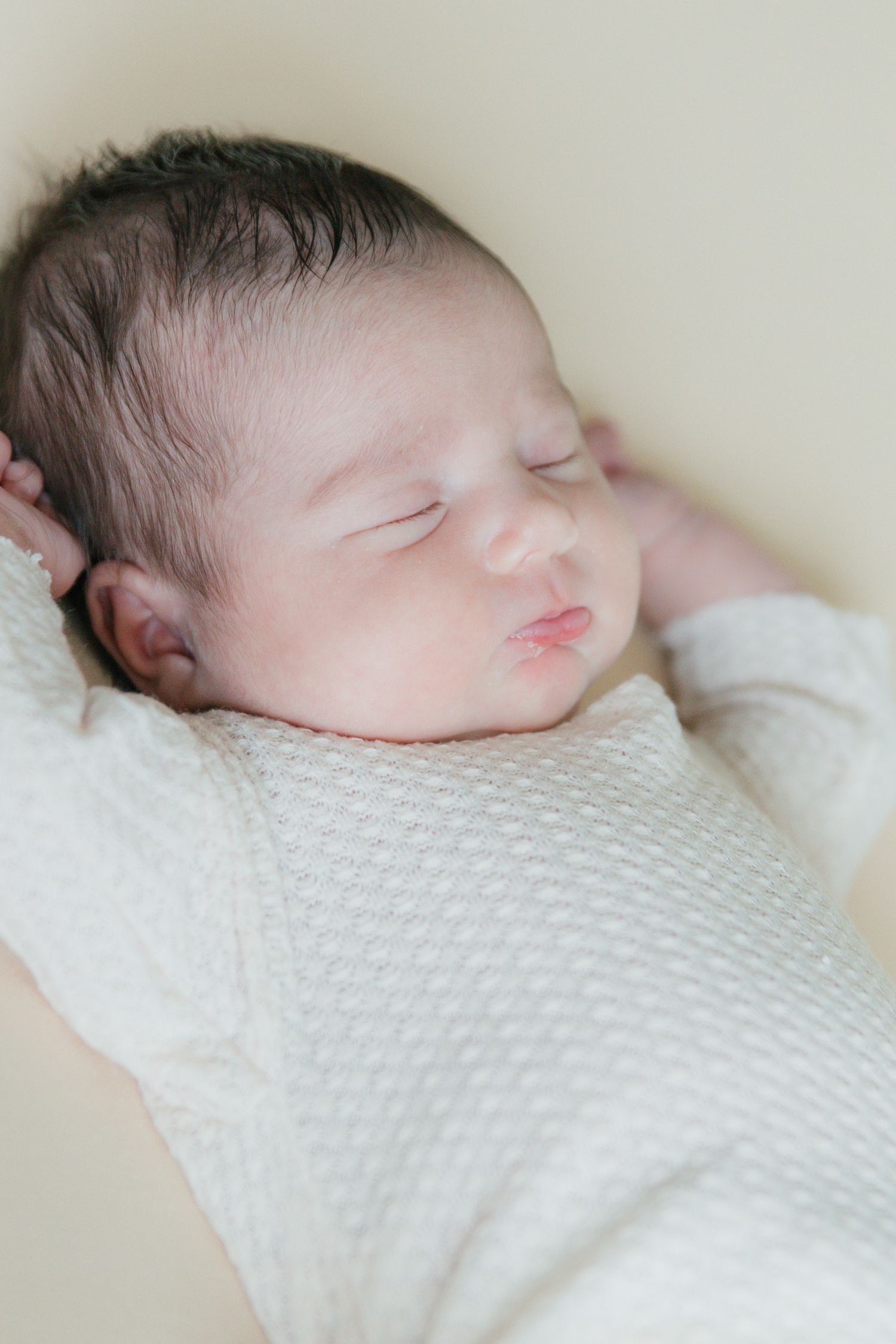 Peaceful sleeping newborn baby in white knit onesie with dark hair and hands raised beside head.