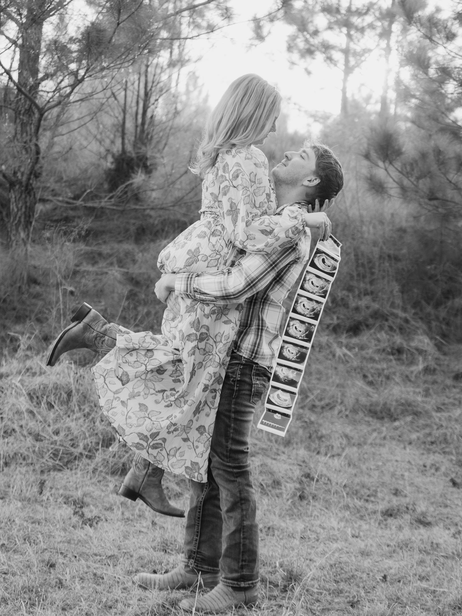 A romantic black and white portrait of a couple embracing in a dreamy forest setting with a floral dress and casual attire.