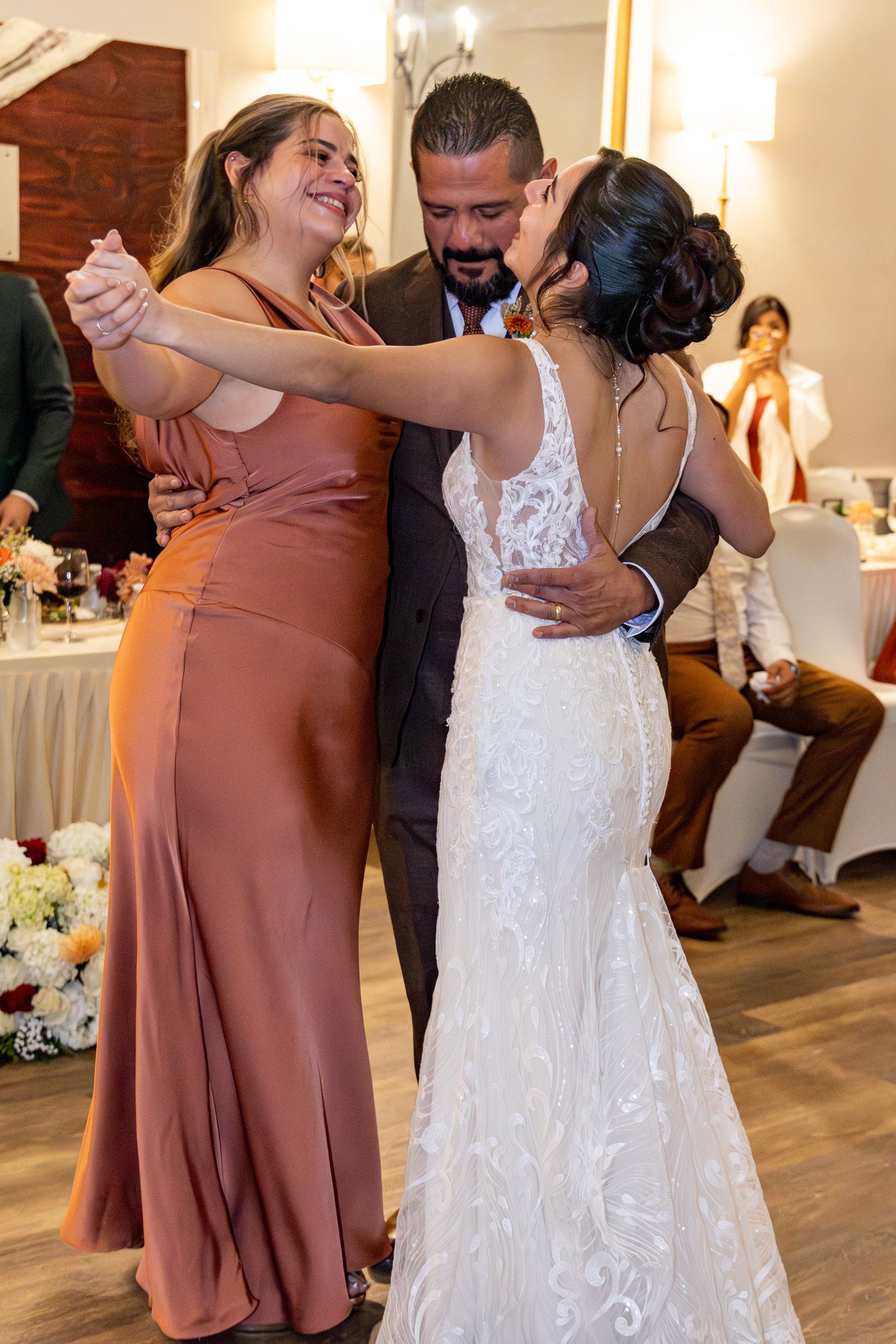 Wedding party members share joyful embraces and dance moves in a reception hall with warm lighting and hardwood floors. Lexington Weddings