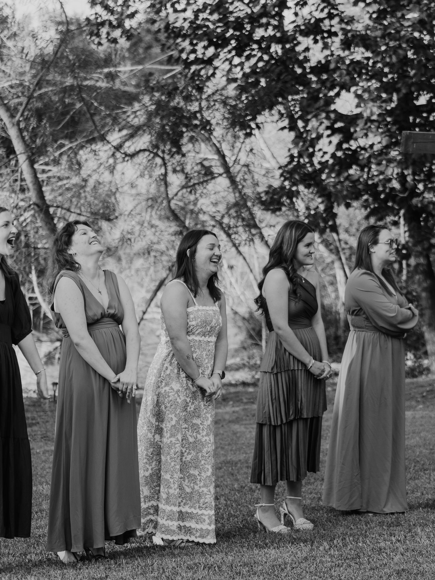Black and white photo of a group of women in long dresses standing together outdoors under trees at a formal event.